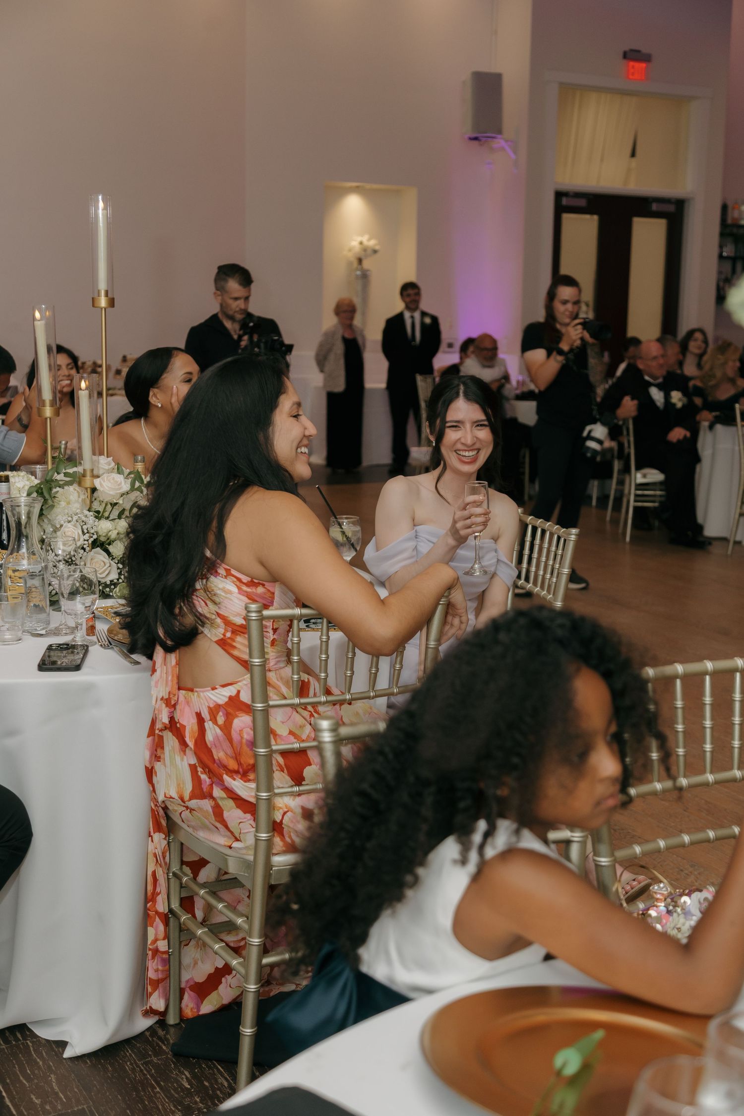 Wedding reception guests laughing and celebrating at elegantly decorated tables in a softly lit ballroom venue.