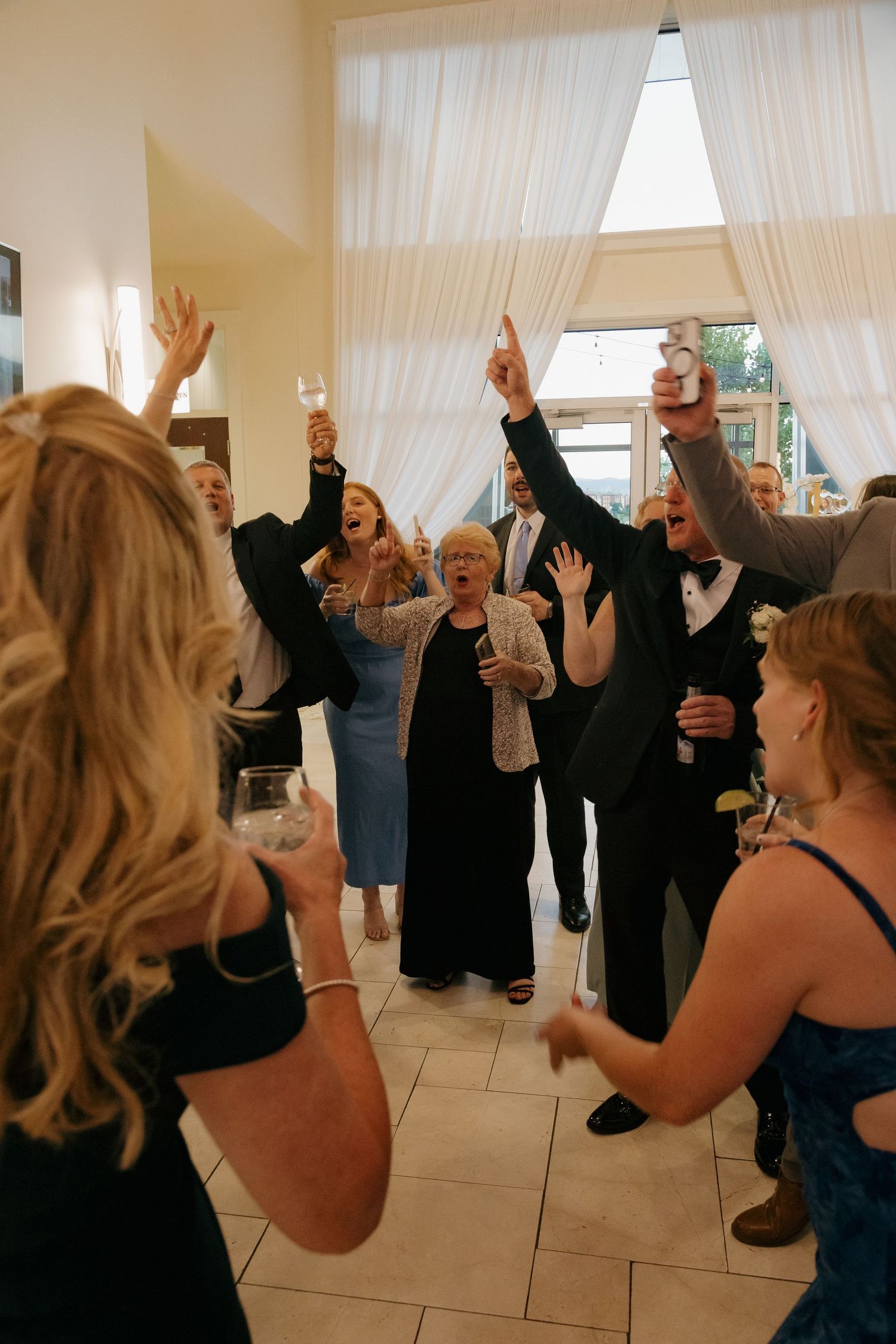 Excited wedding guests raise hands and glasses in celebration during lively indoor reception dance.