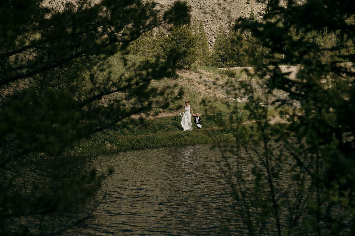 Bride in white dress standing by a serene mountain lake surrounded by lush pine trees and rocky cliffs.