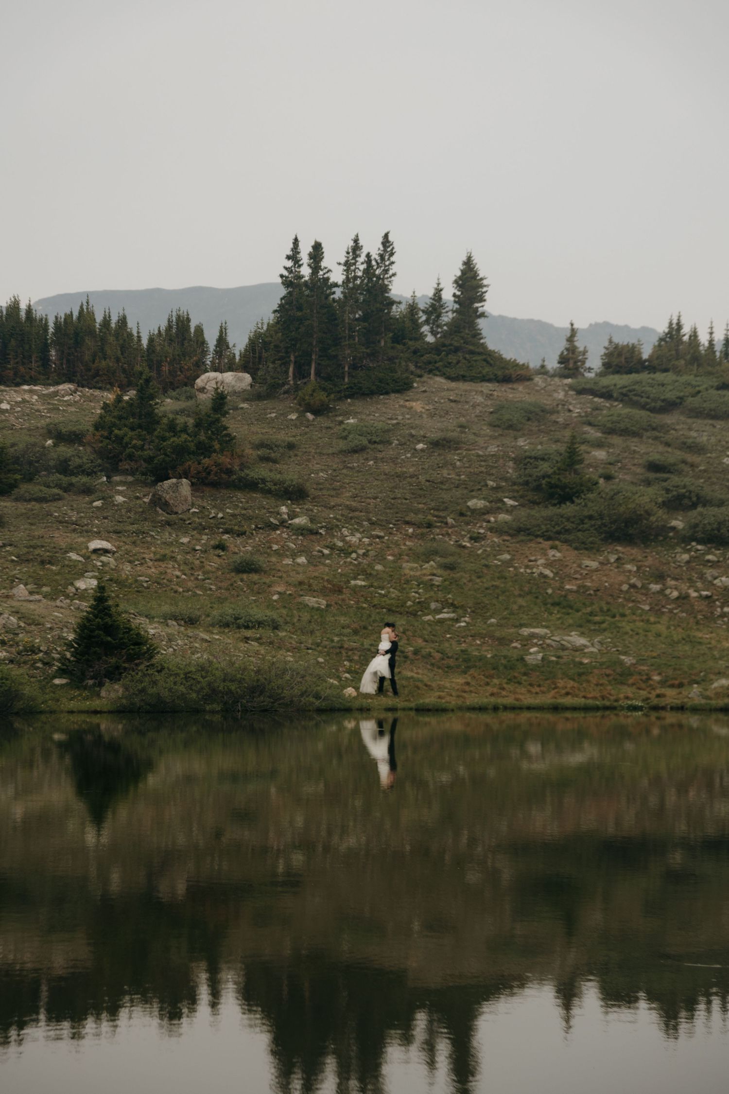 Wedding couple walks hand in hand beside a serene mountain lake reflecting evergreen trees and hills.