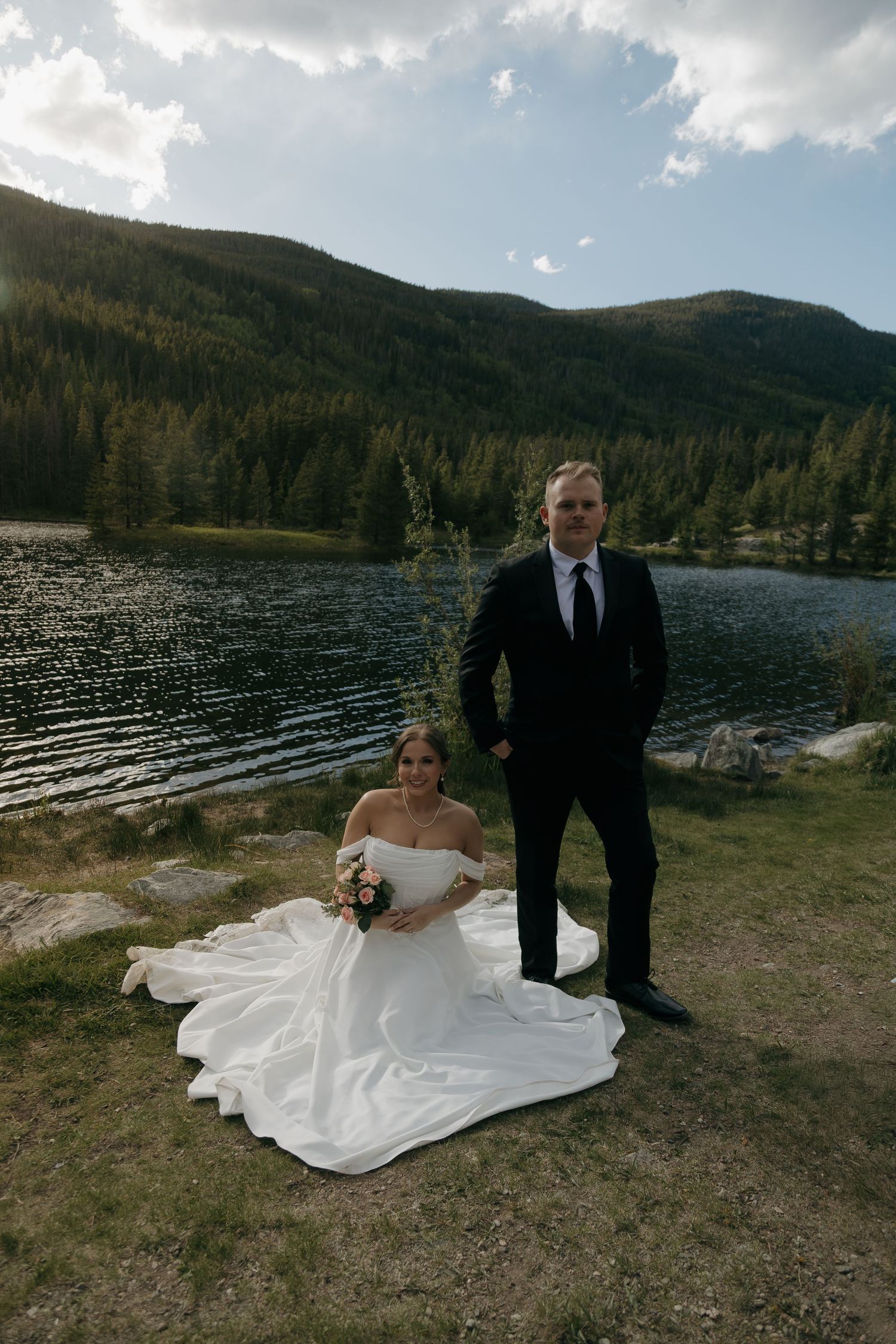 Bride sits on grass with flowing gown as groom stands beside her near a scenic mountain lake.