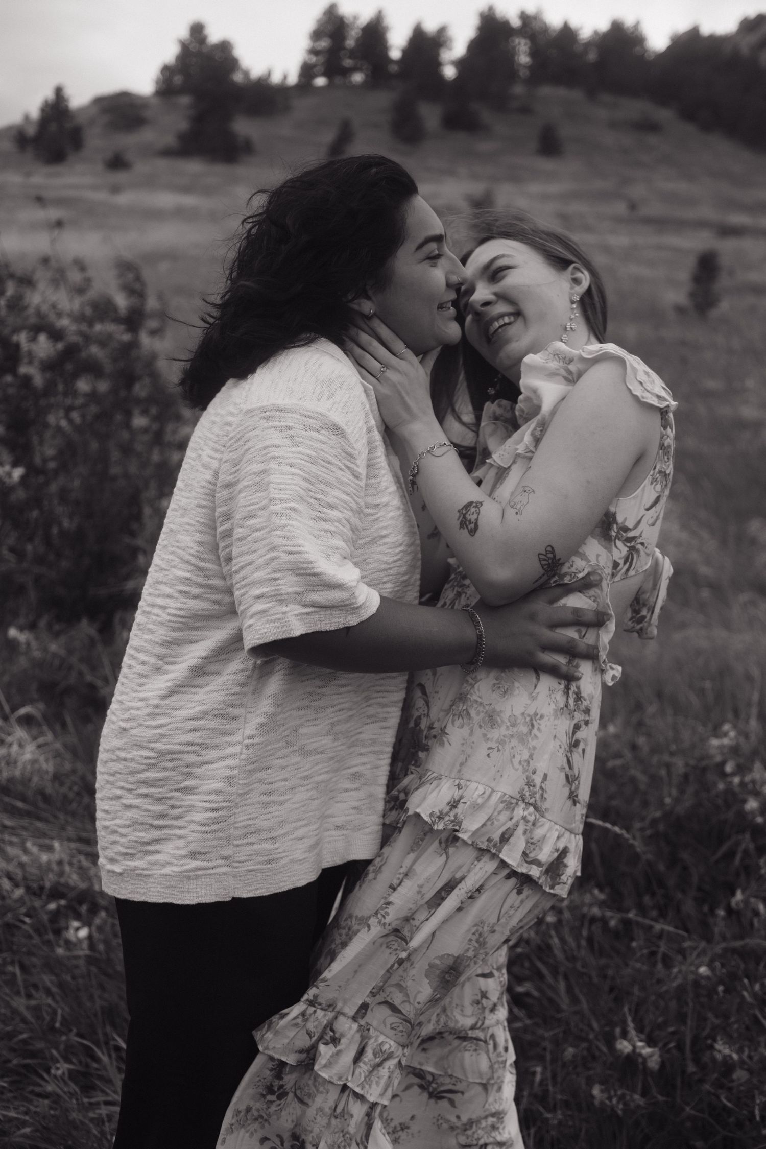 Black and white photo of two women laughing and embracing in a grassy outdoor landscape.