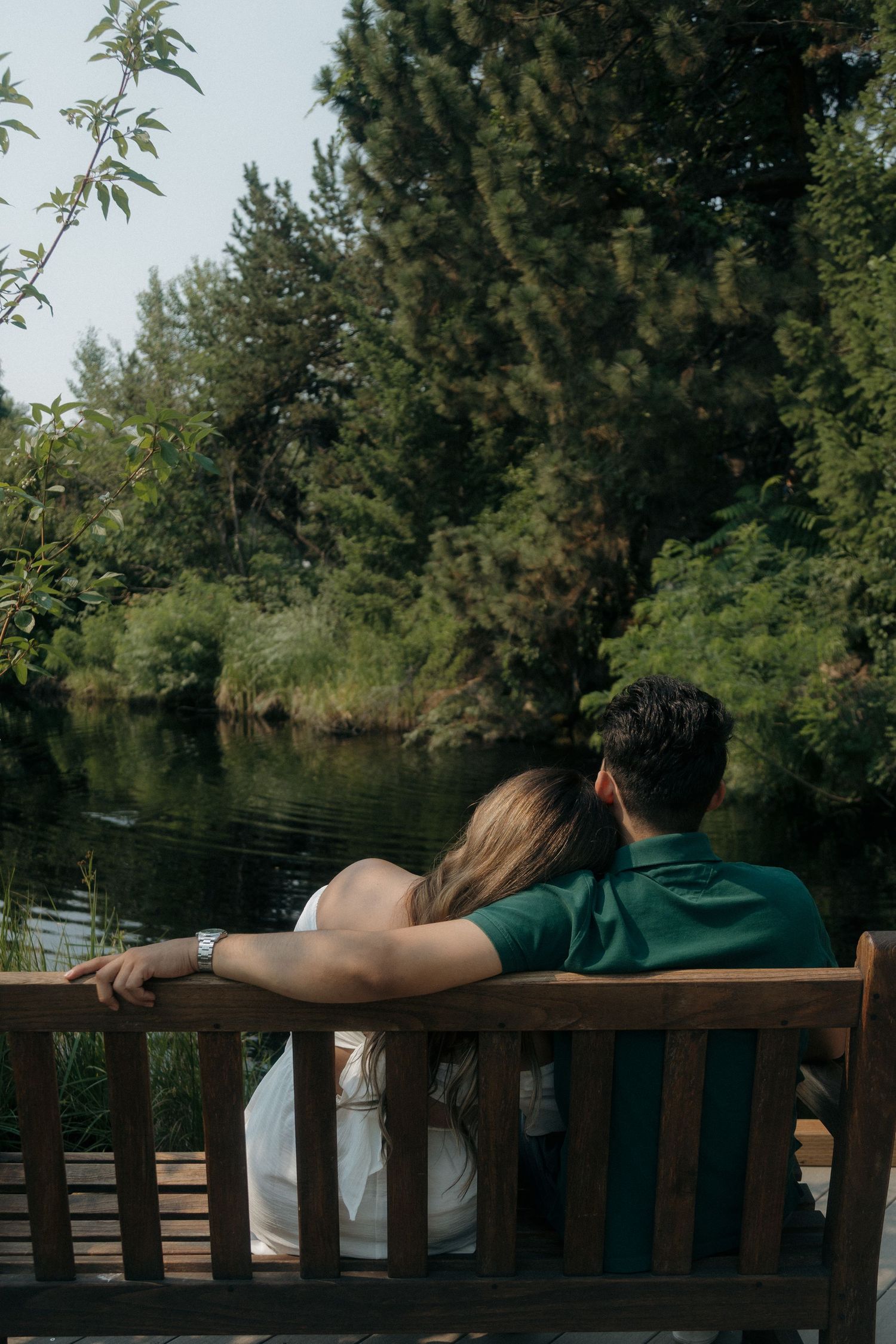 Couple sitting together on a wooden bench overlooking a serene pond surrounded by tall green trees.