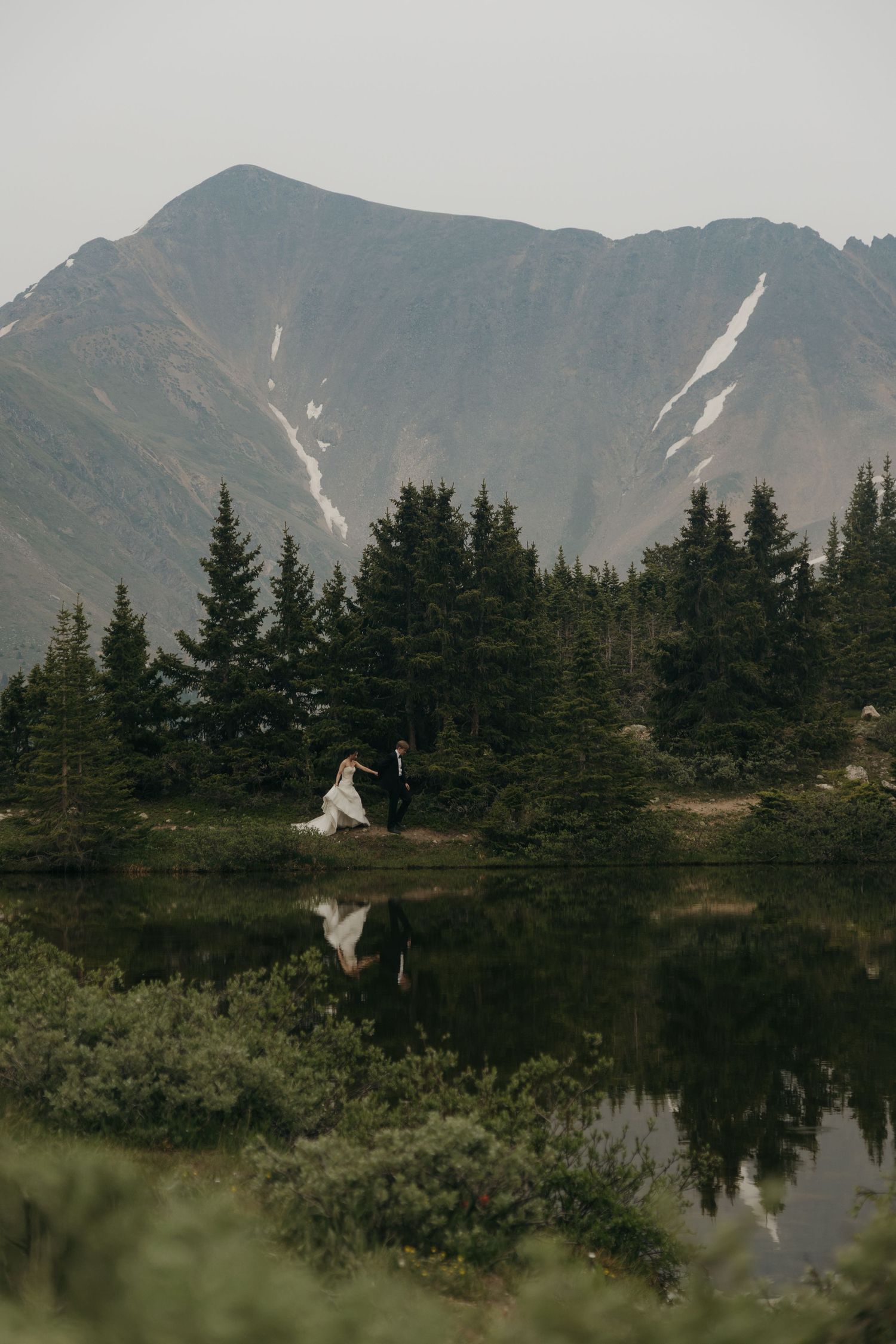 Bride and groom walk hand in hand beside a reflective alpine lake with evergreen trees and a misty mountain backdrop.