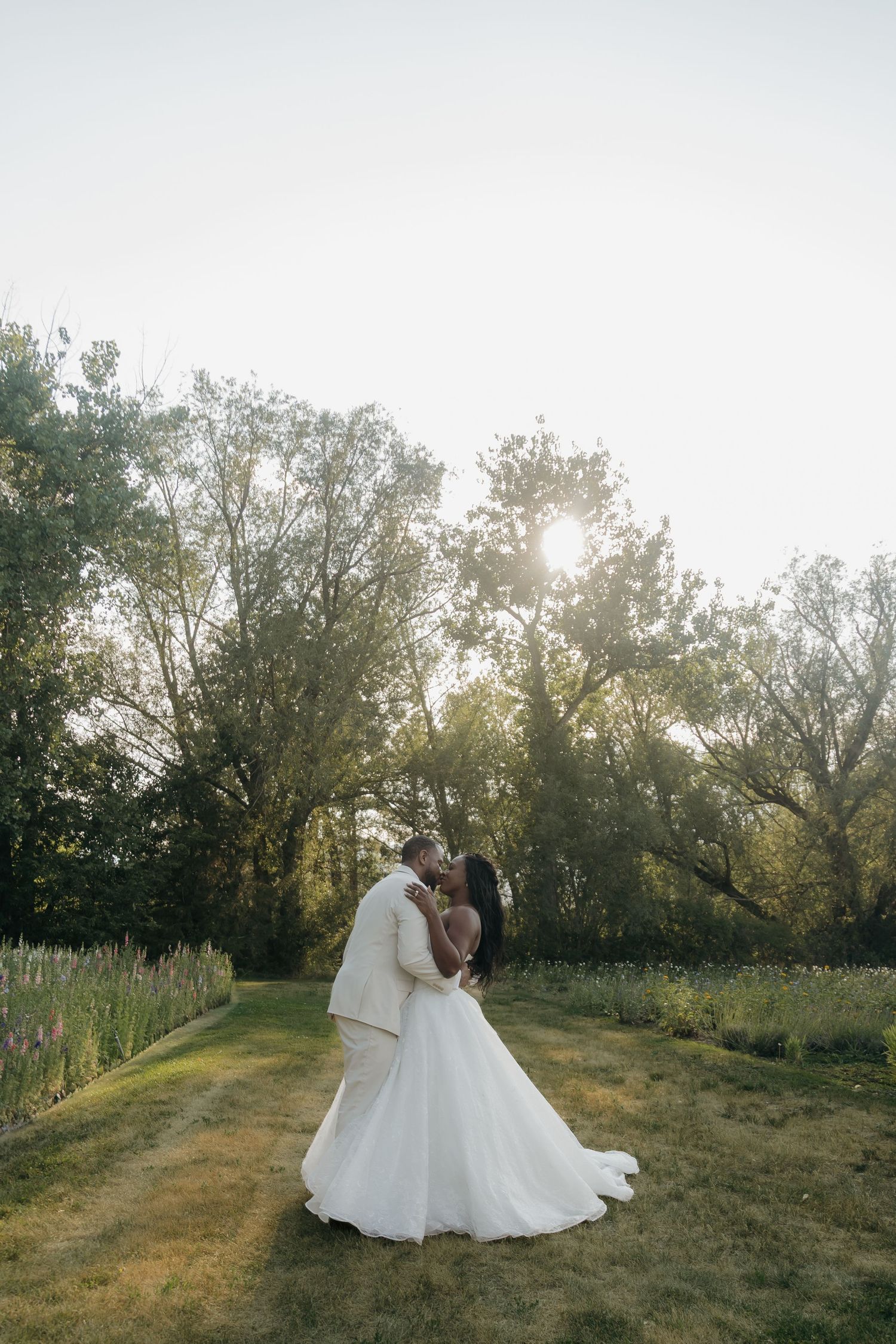 Bride and groom share a romantic kiss at golden hour in a lush garden setting, with sunlight filtering through tall trees.