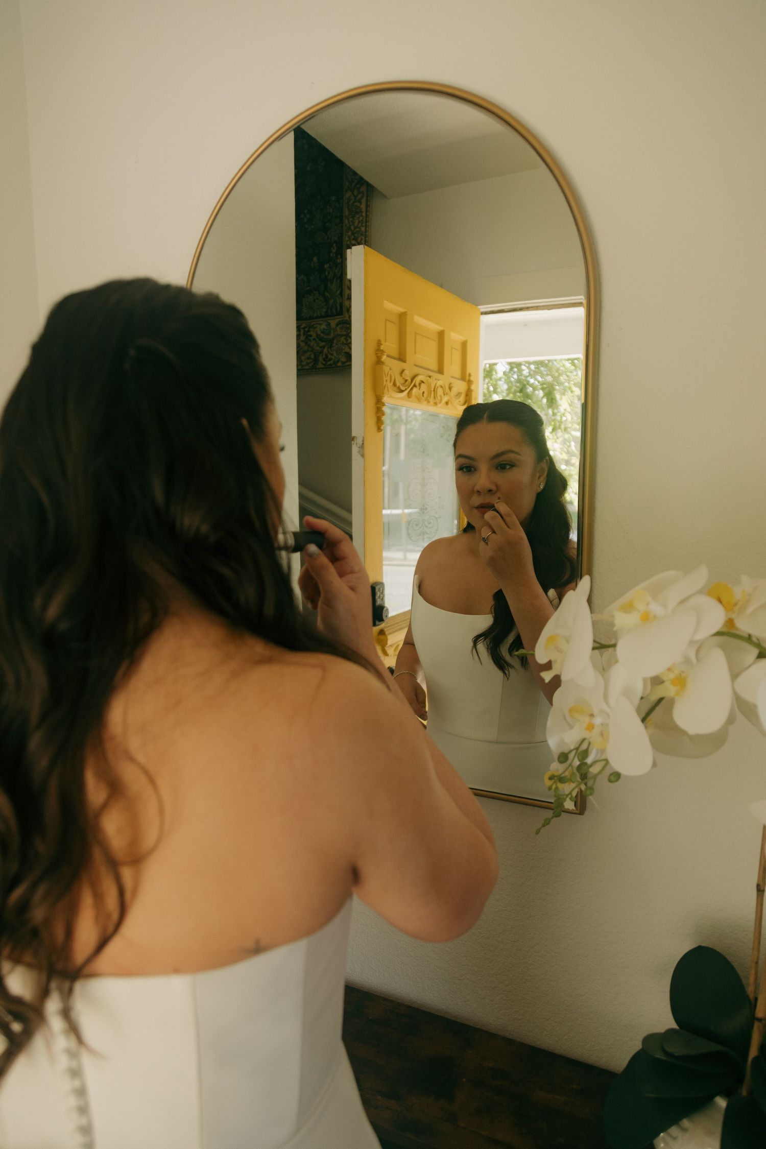 Bride applying makeup while looking in an oval gold-framed mirror on her wedding day.
