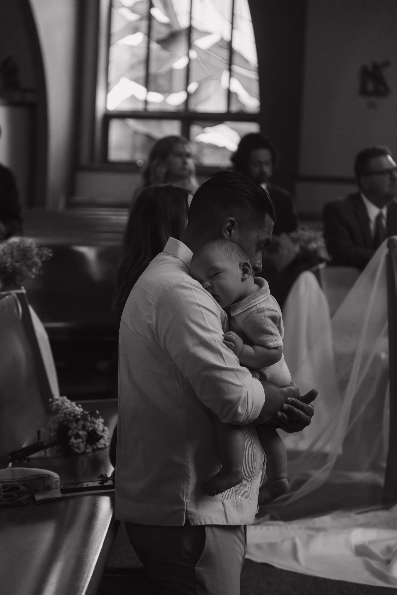 A man tenderly holds a sleeping baby during a church ceremony, with guests seated in pews behind.