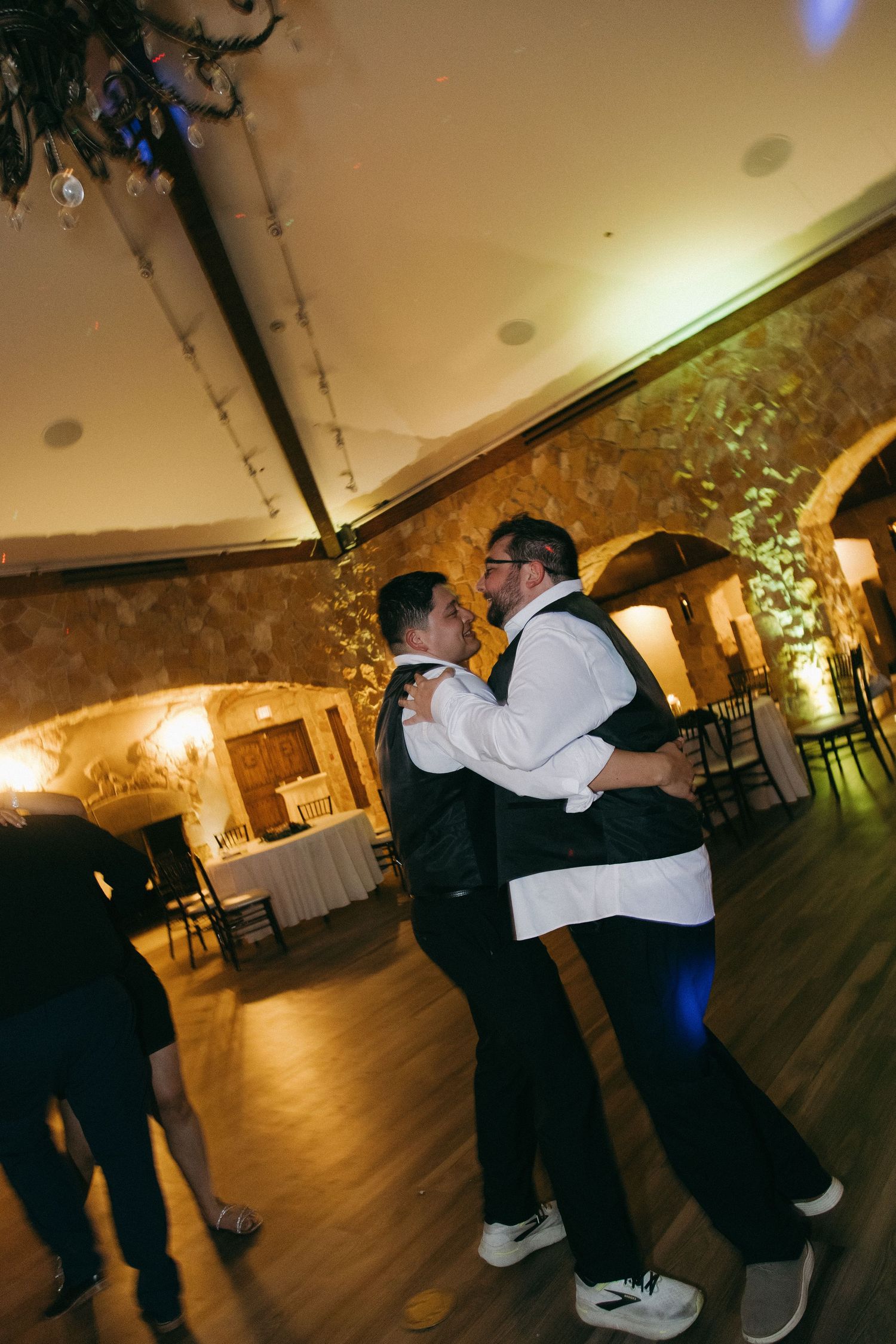 Groom dancing with groomsman on wooden dance floor in warmly lit stone-walled wedding venue.