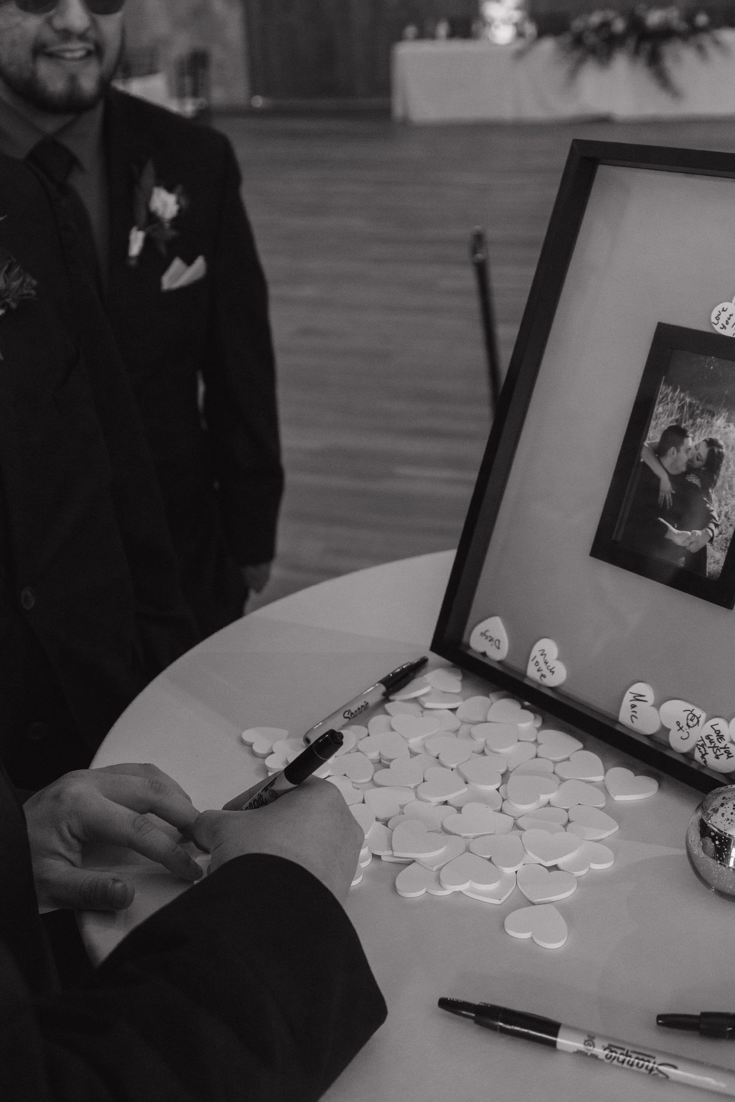 Black and white photo of wedding guest signing heart-shaped cards in a shadow box guest book at reception.