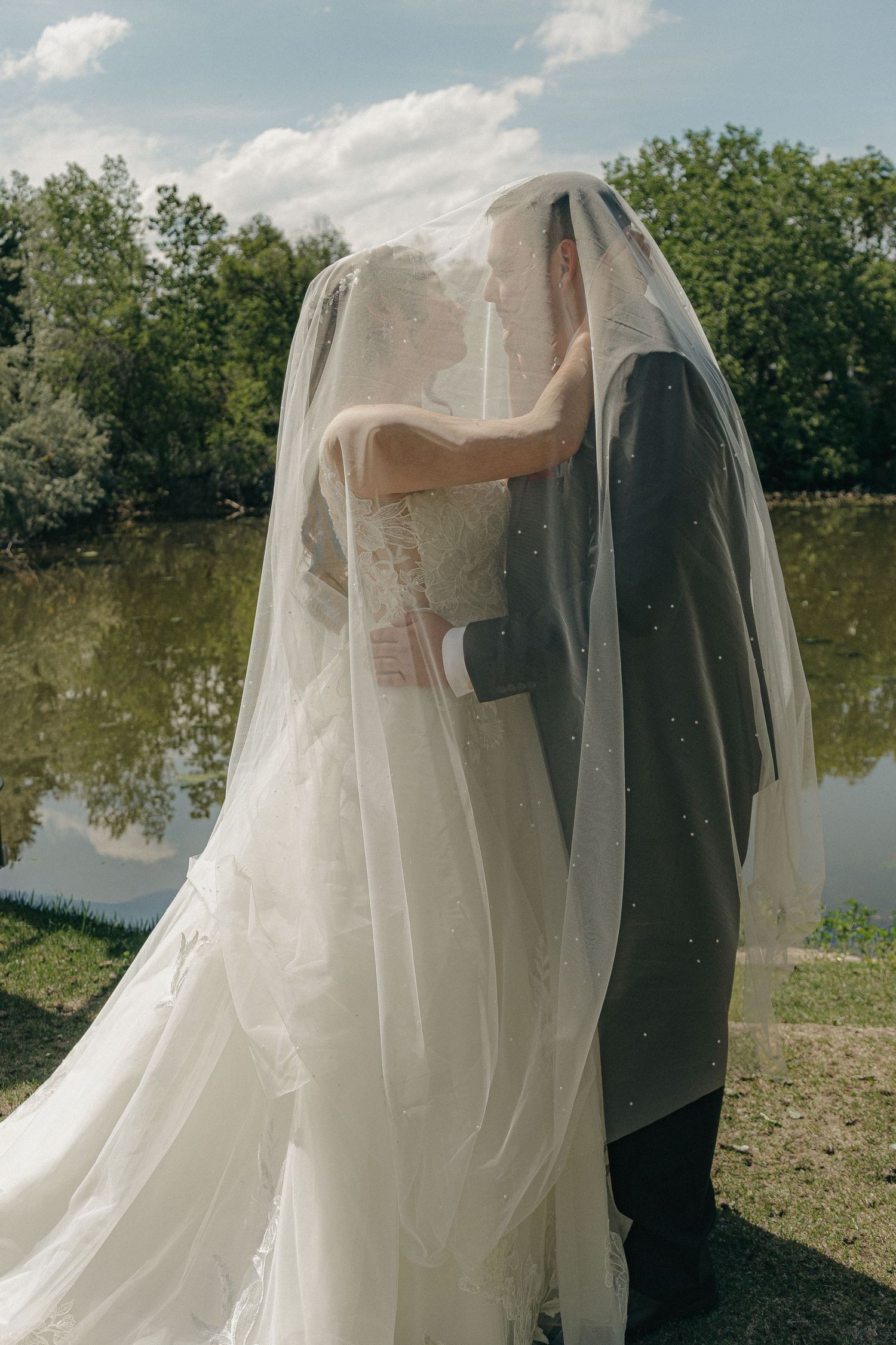 Bride and groom hidden under flowing white veil share intimate moment by a scenic lake.