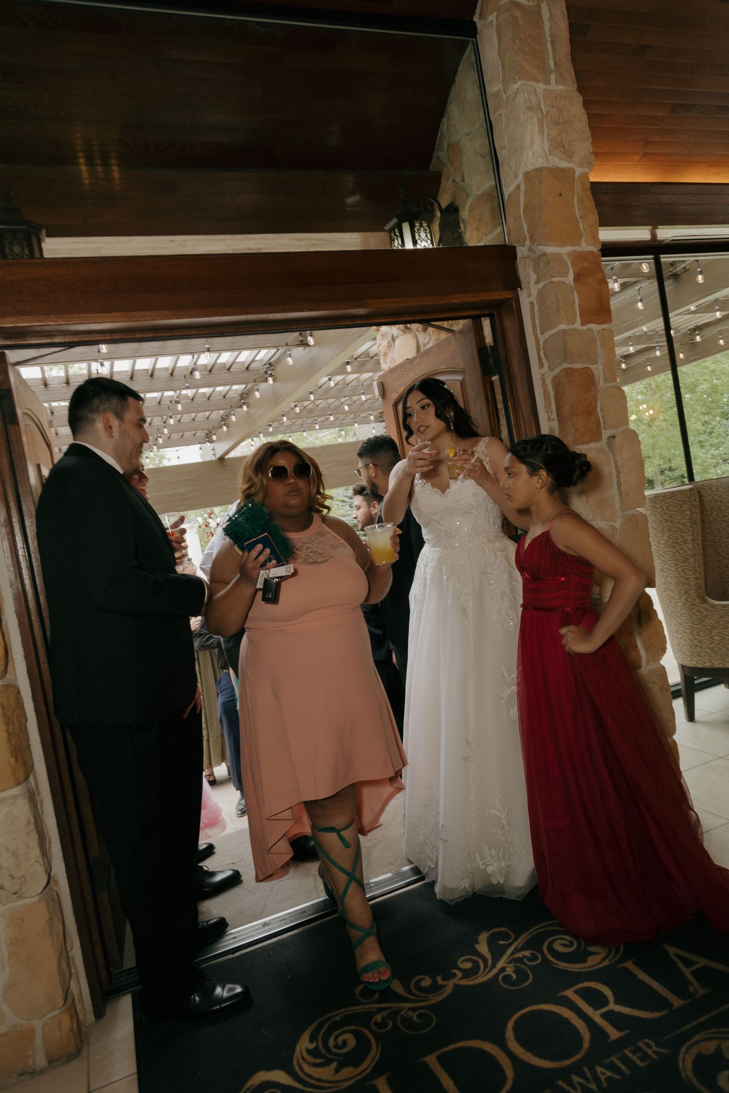 Wedding guests in formal attire gather at venue entrance, bride in white gown holds champagne glass.