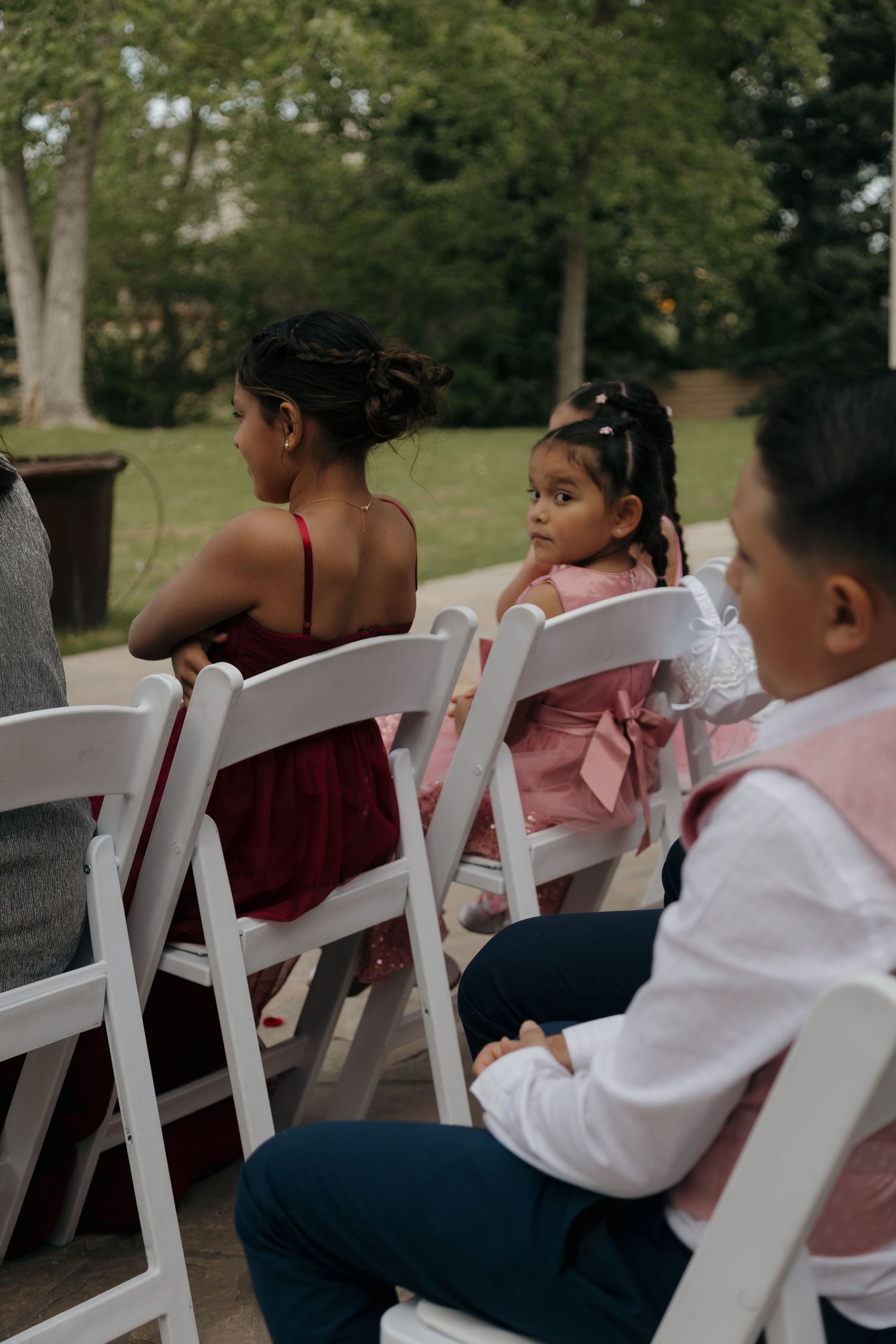 Children in formal attire seated in white chairs at an outdoor wedding ceremony.