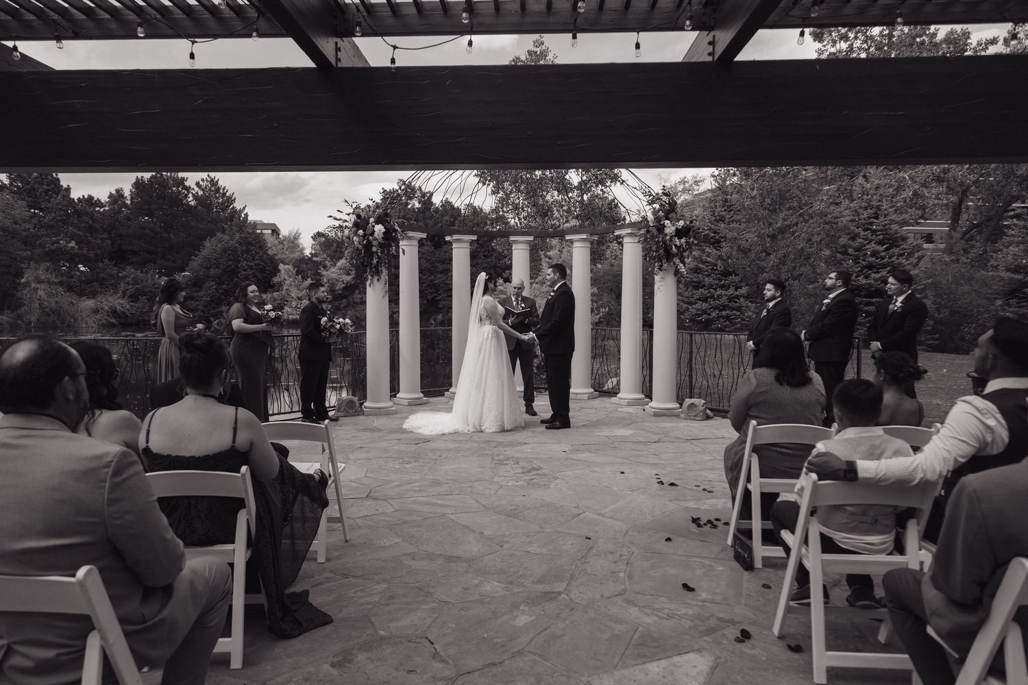 Black and white wide shot of outdoor wedding ceremony with guests seated in white chairs.