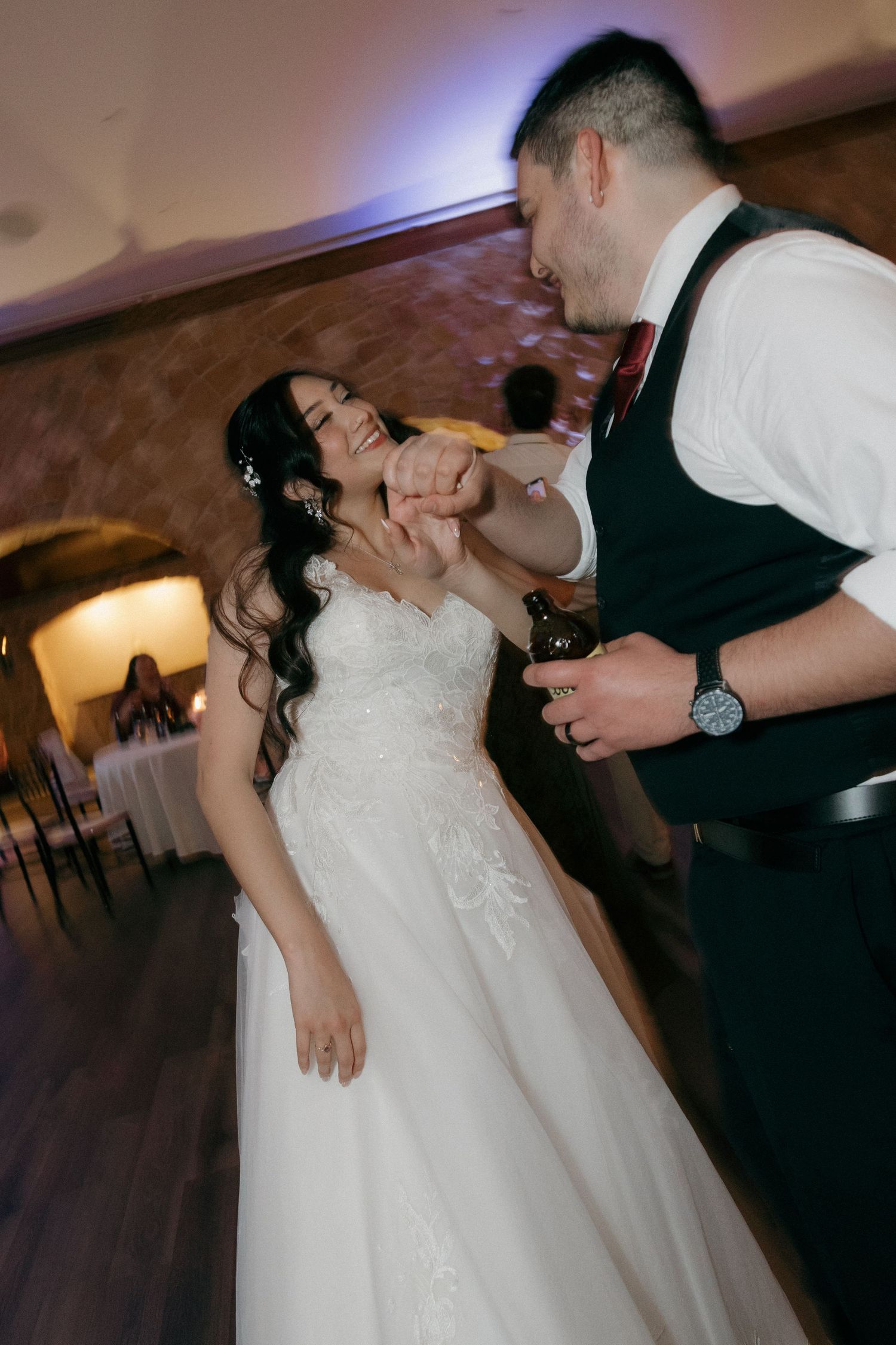 Laughing bride in white gown dancing with groom in vest at wedding reception.