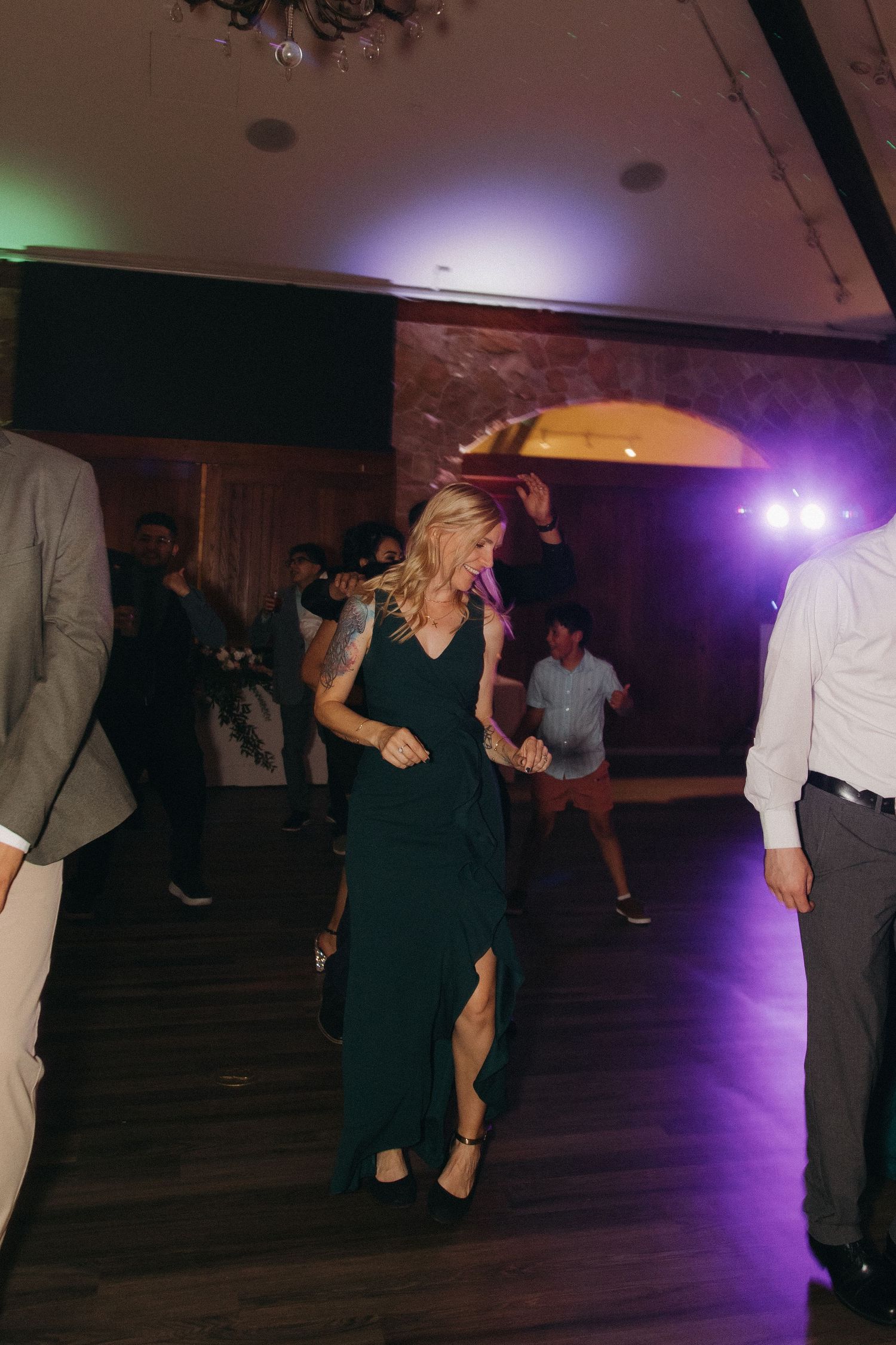 Blonde woman in green dress dancing energetically at wedding reception with purple stage lighting.