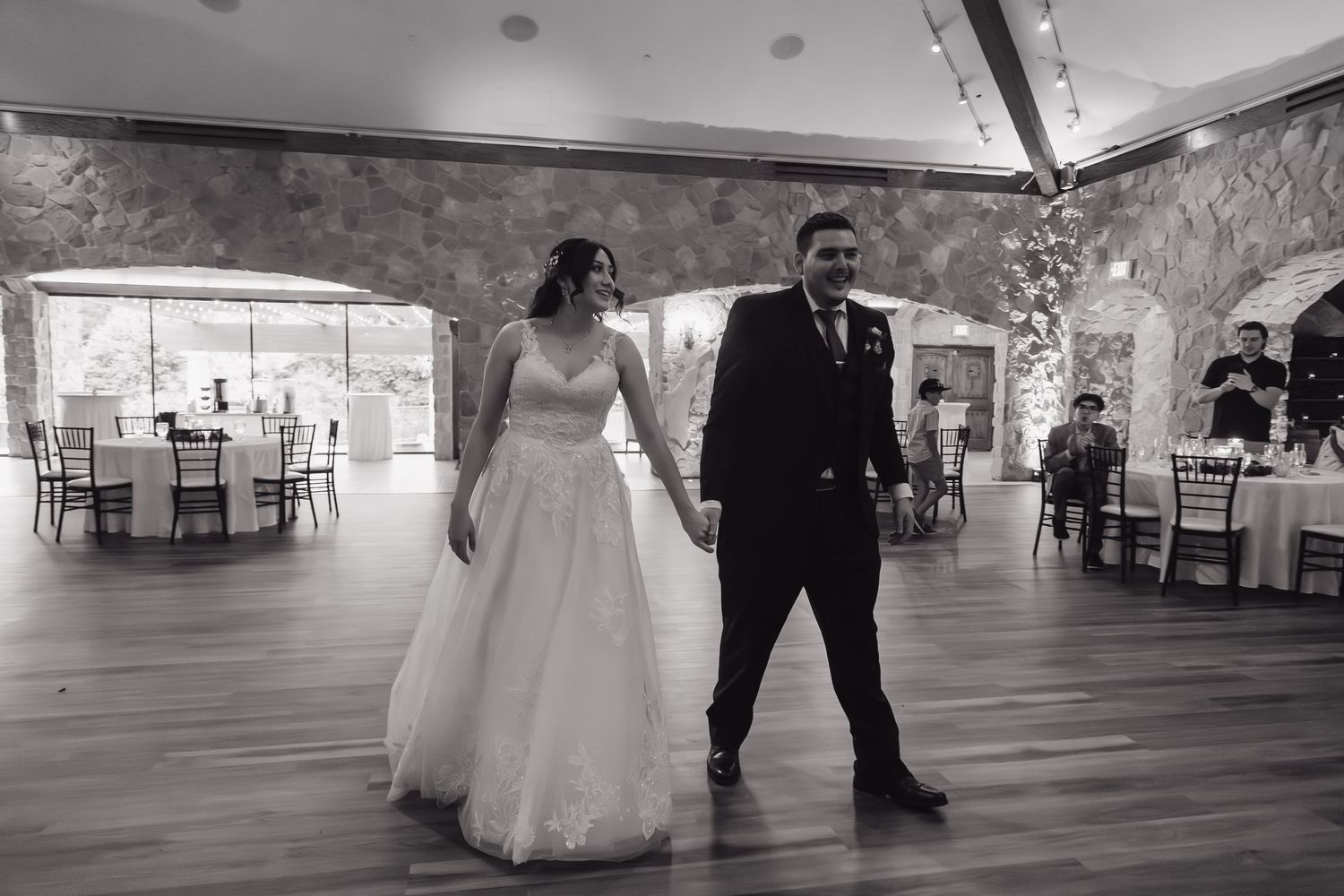 Black and white photo of bride and groom walking onto the dance floor in a stone-walled reception hall.