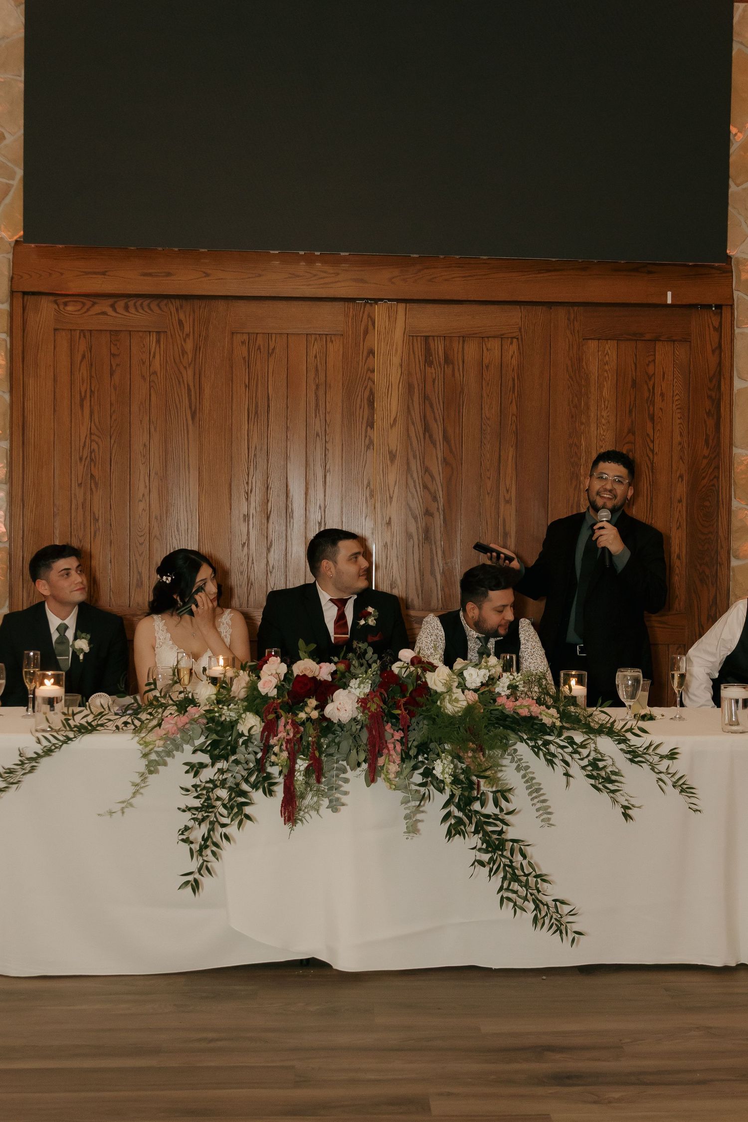 Wedding party seated at the head table during a reception toast with floral arrangements.