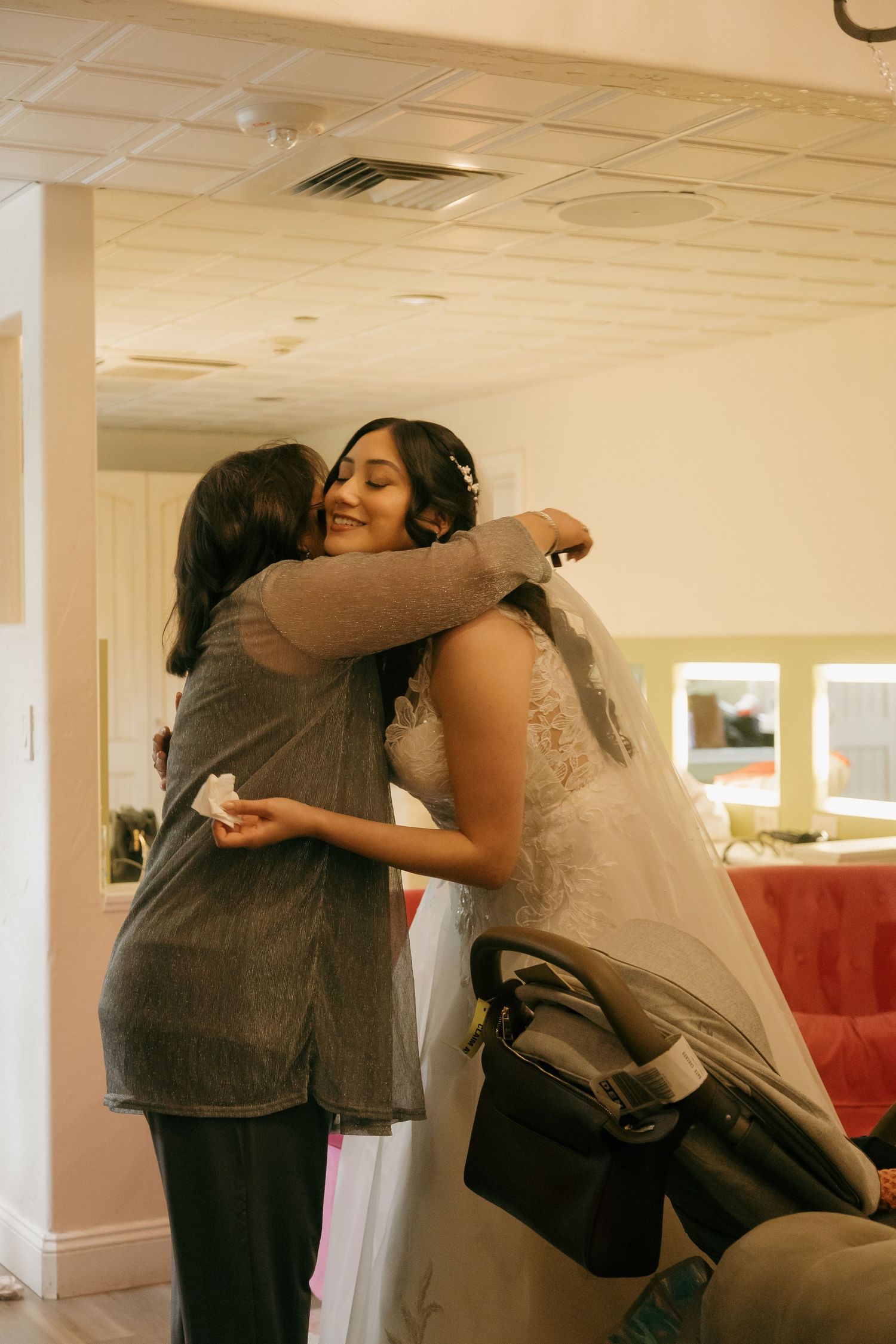 Bride in white lace gown and veil shares an emotional hug with a guest in a bridal suite.