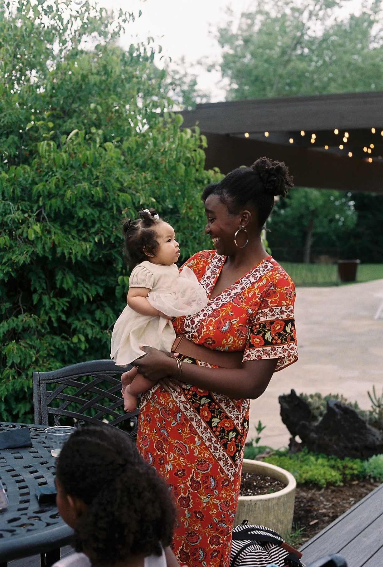 Elegantly dressed woman in floral gown holds a baby at an outdoor wedding reception garden.