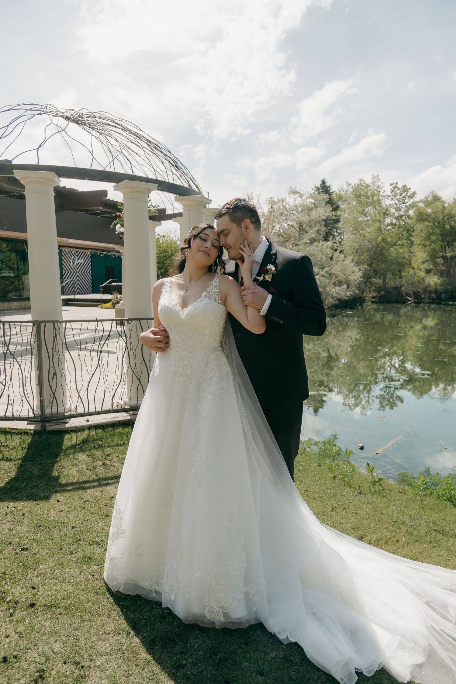 Groom tenderly kisses bride near a decorative gazebo by a reflective pond on their wedding day.