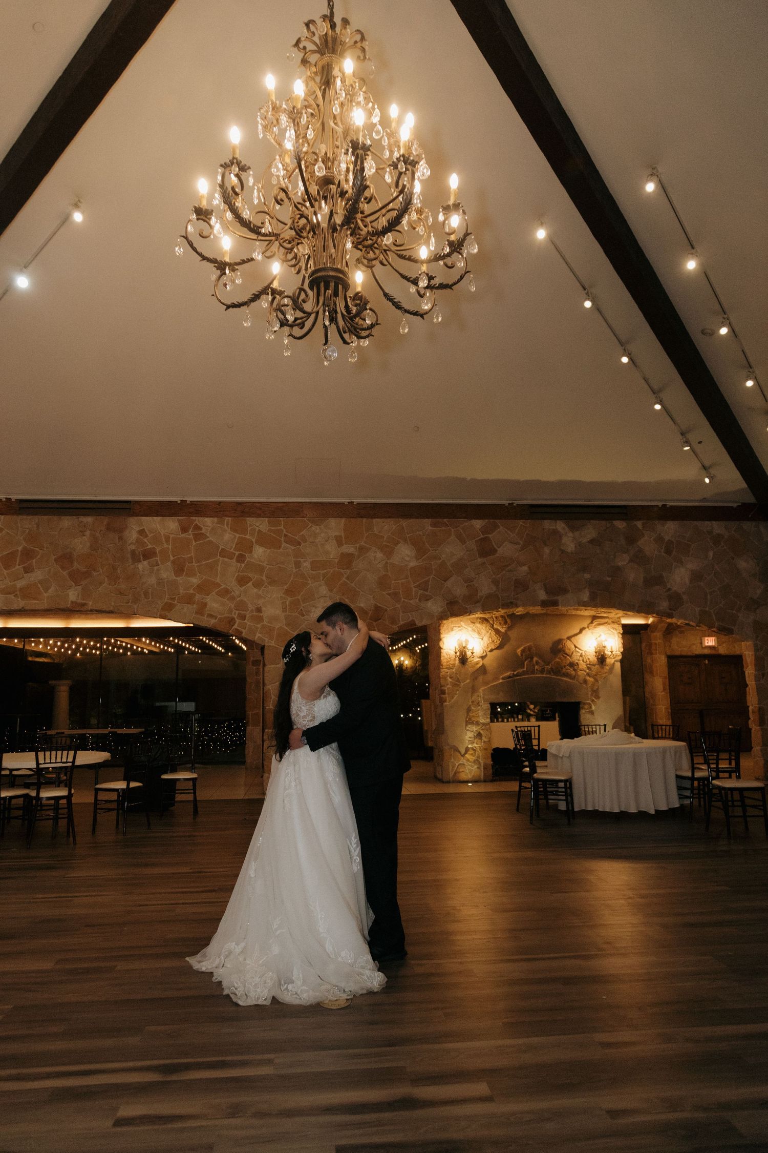 Bride and groom share a last dance under an elegant chandelier in a rustic stone wedding ballroom.