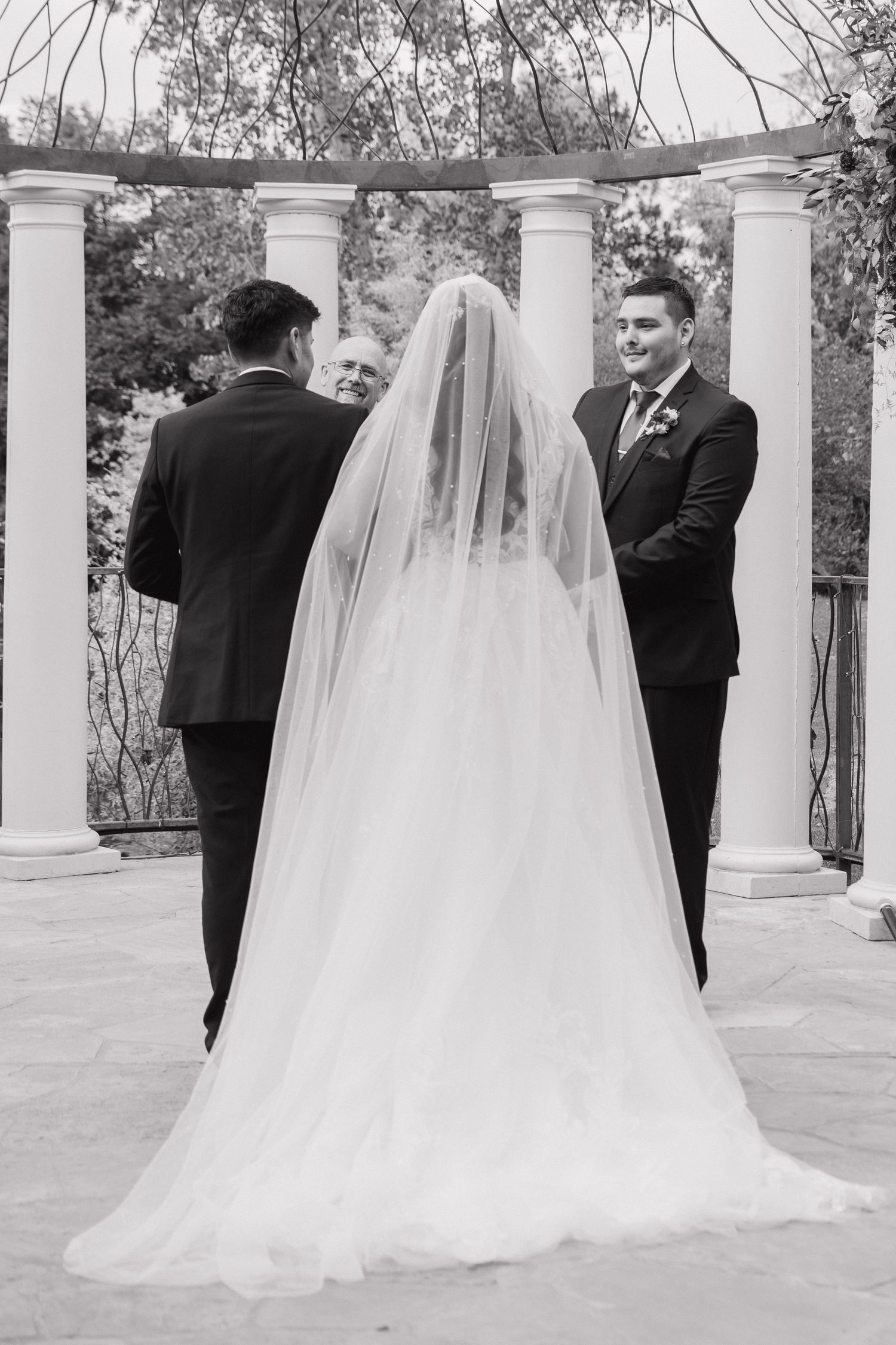 Black and white wedding ceremony photo of bride in veil and groom standing before officiant at columned outdoor altar.
