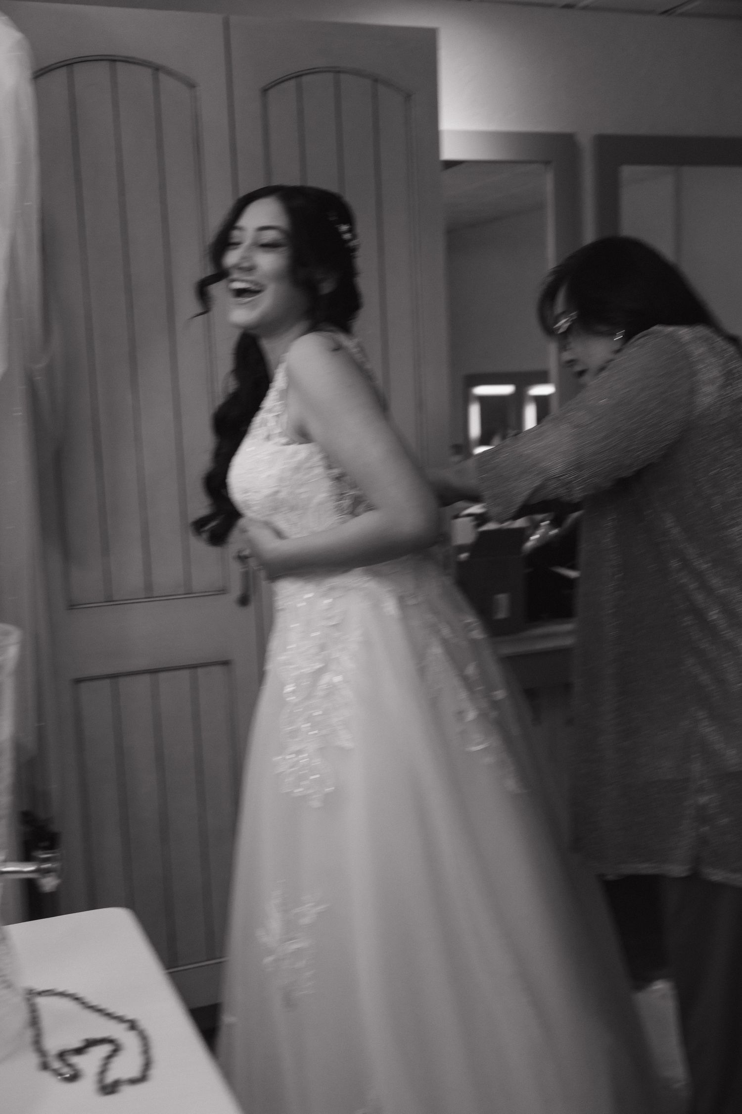Bride laughing in white lace wedding gown while assistant helps fasten dress in black and white photo.