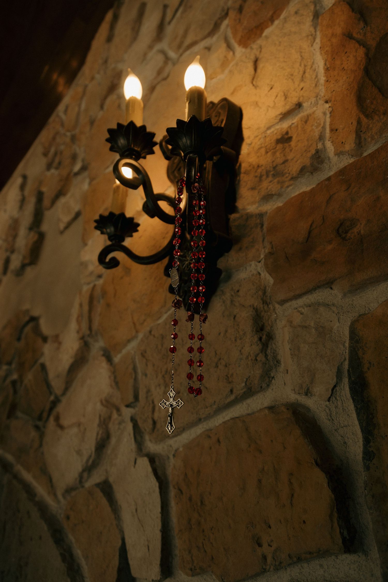 Ornate wrought iron wall sconce with lit candles and red rosary beads on a stone wall.