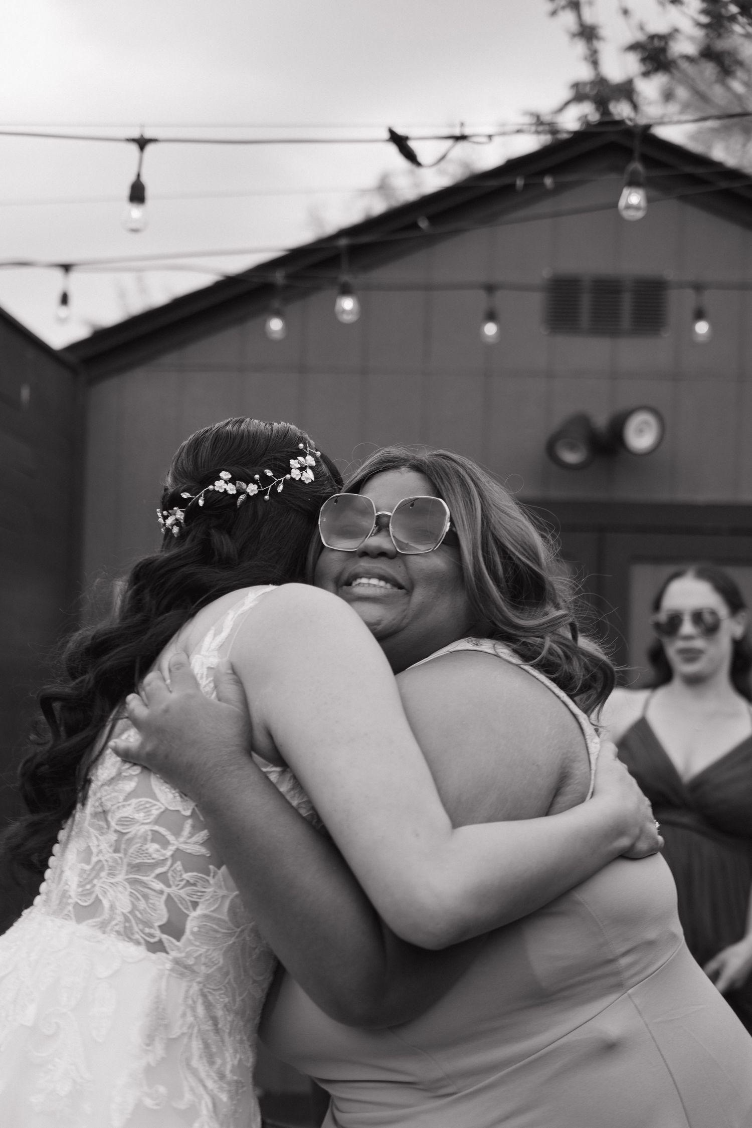 Black and white photo of bride in lace gown sharing a warm hug with a smiling guest at wedding.