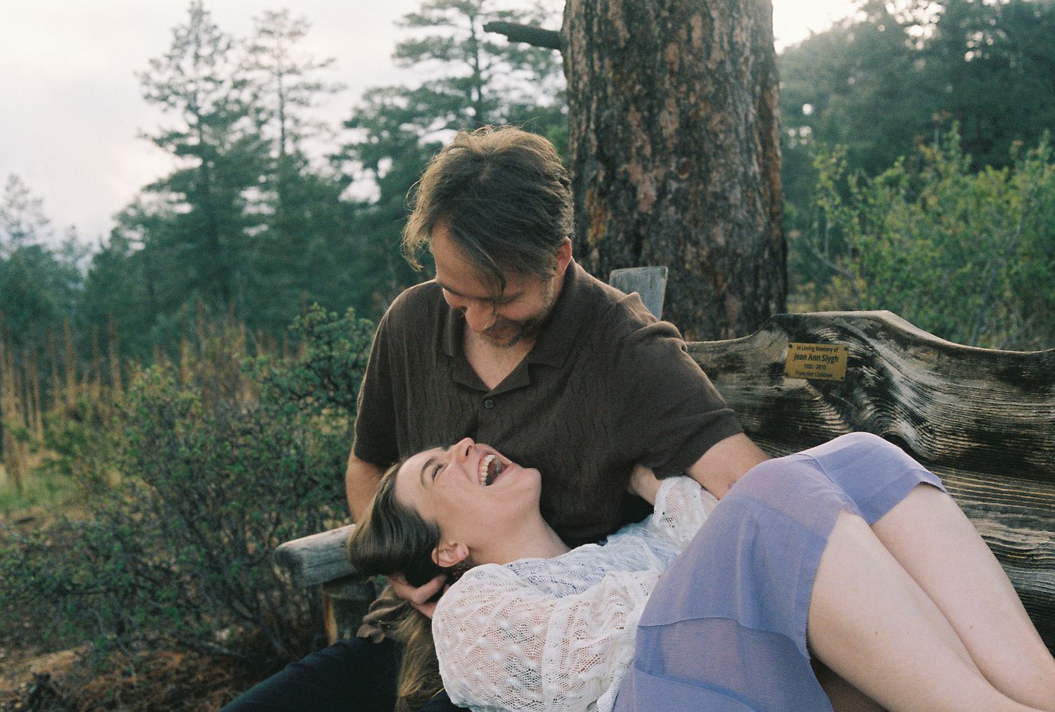 Couple sharing a tender moment near a rustic log, woman laughing while lying back in a forest setting.