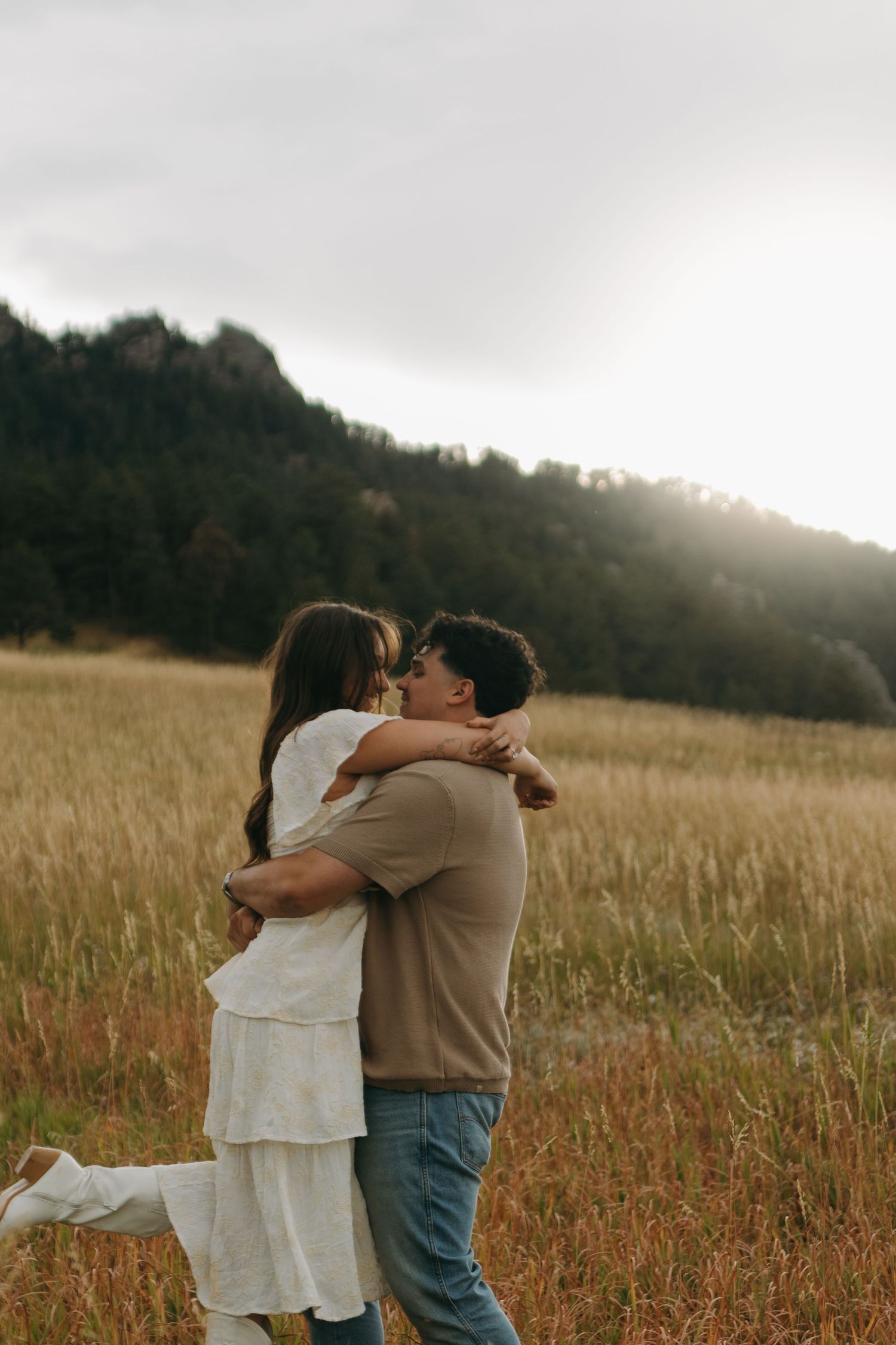 Couple embracing and almost kissing in a sunlit golden meadow with mountain scenery.