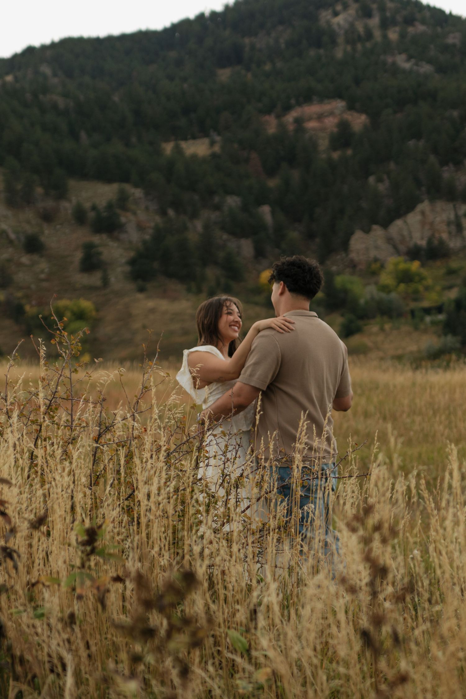 Couple embracing and laughing together in tall golden grass field with pine covered mountain backdrop.