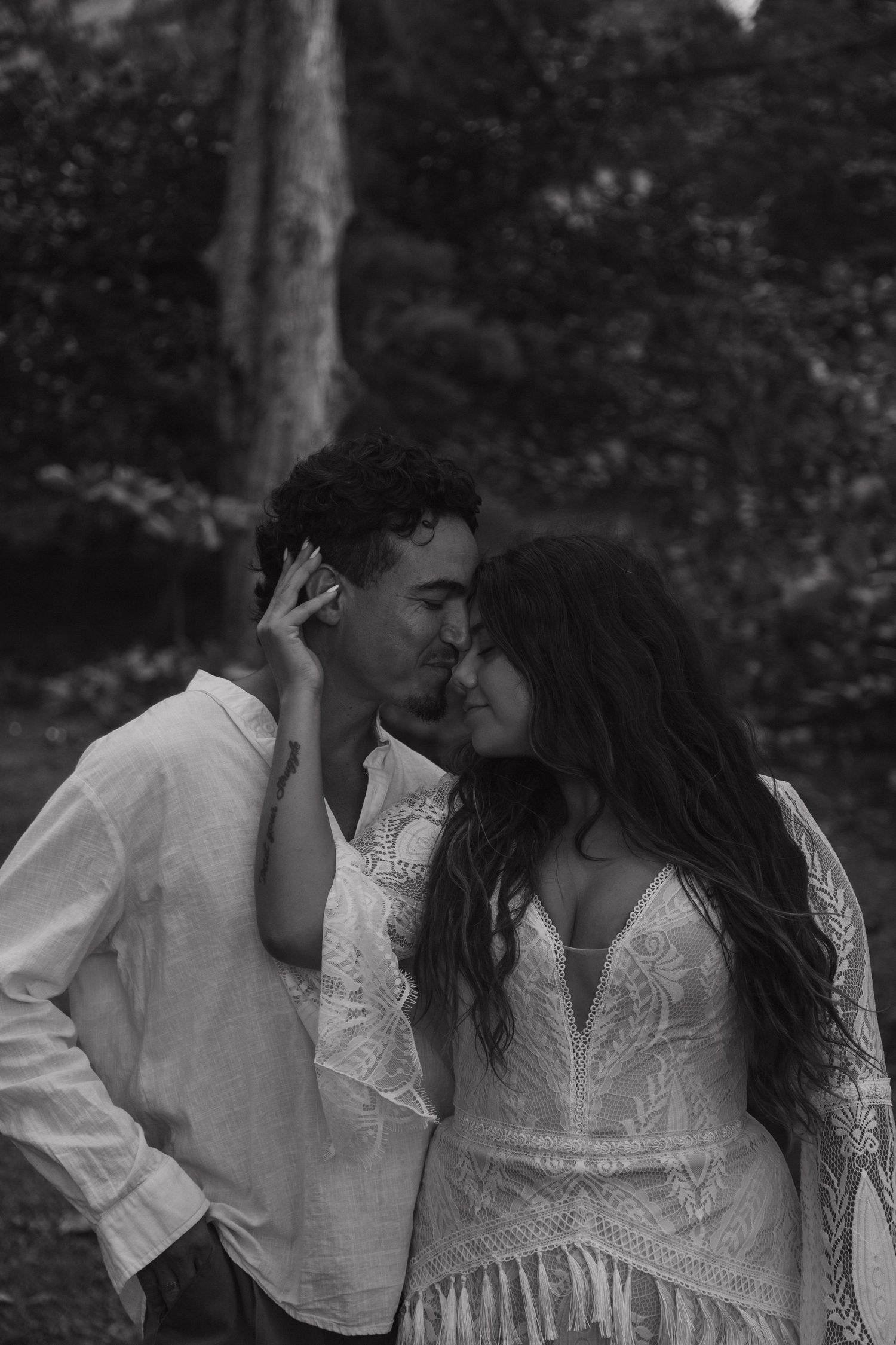 Romantic black and white portrait of couple nearly kissing, woman in boho lace dress touching mans face outdoors.