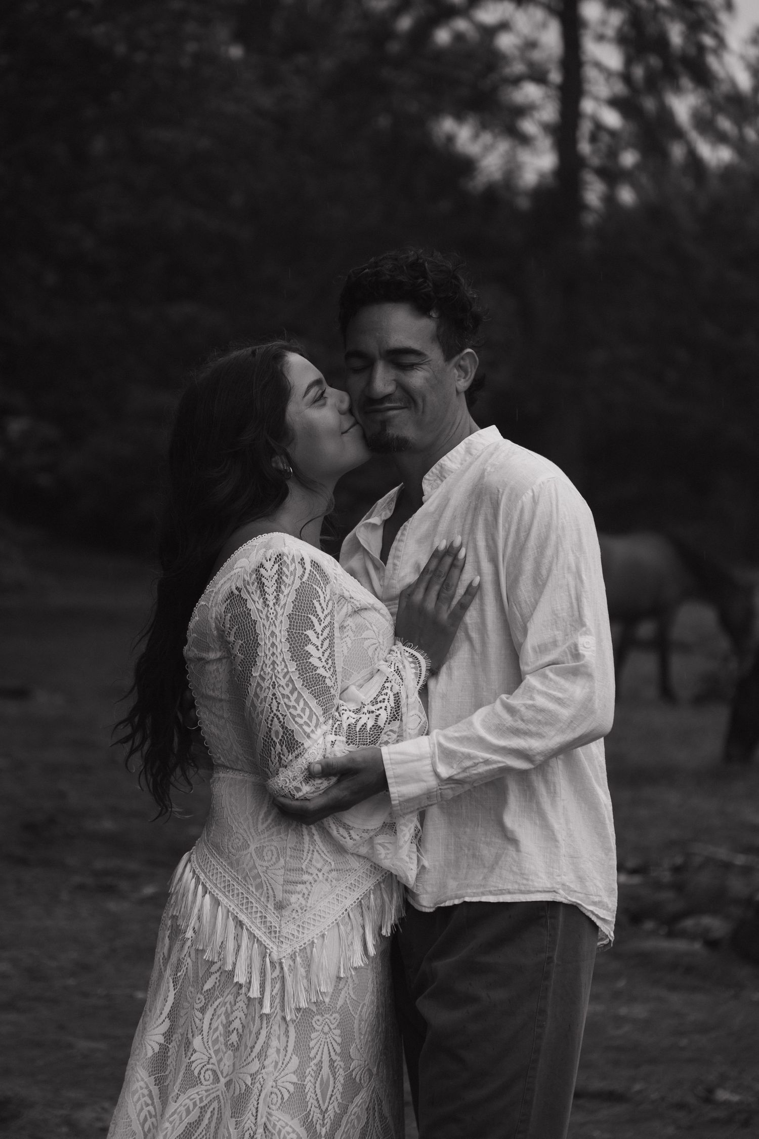 Black and white photo of a couple embracing outdoors, woman in lace dress kissing mans cheek while he smiles.