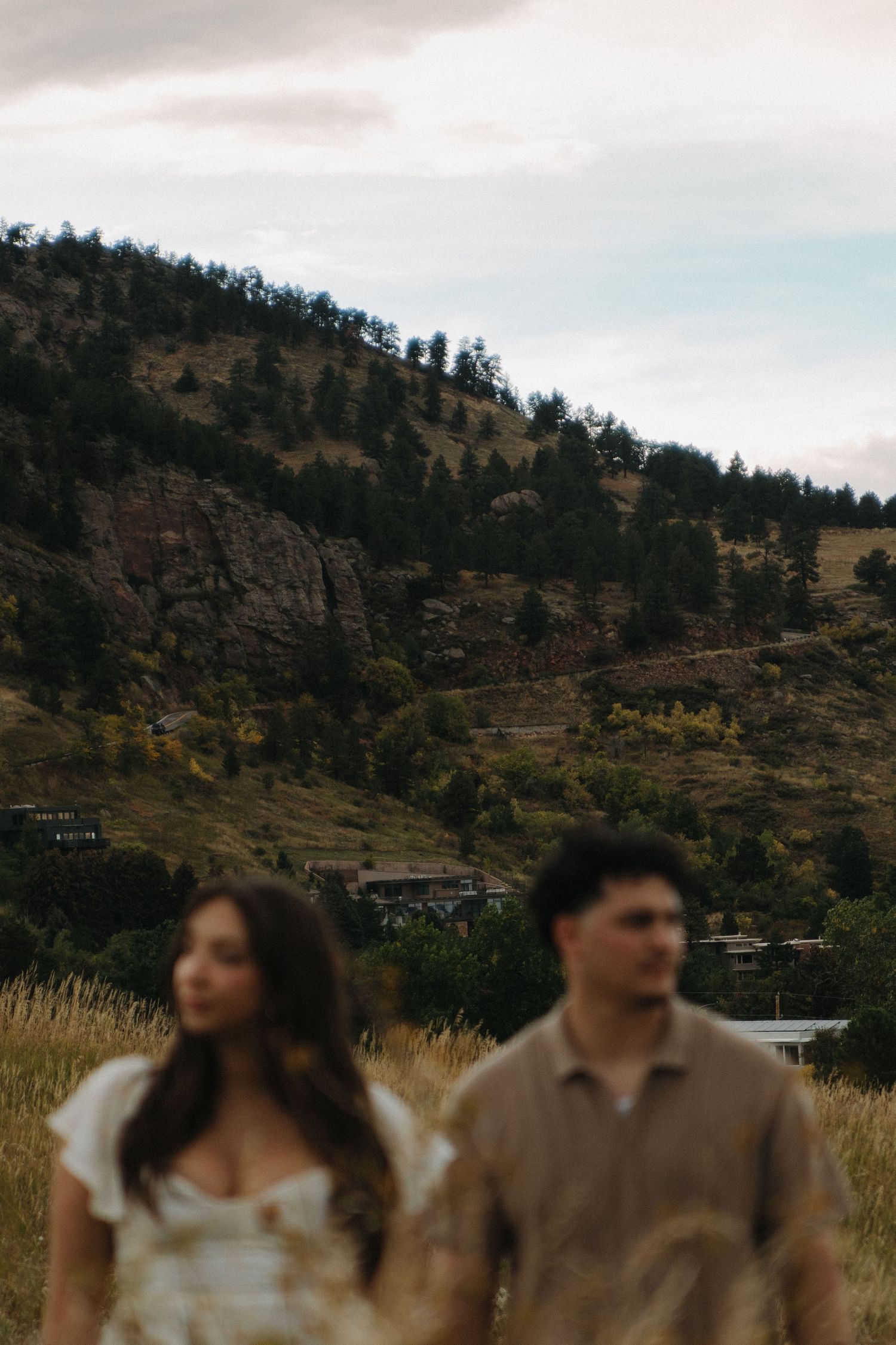 Couple walks through golden field with mountain backdrop during outdoor engagement session in Colorado.