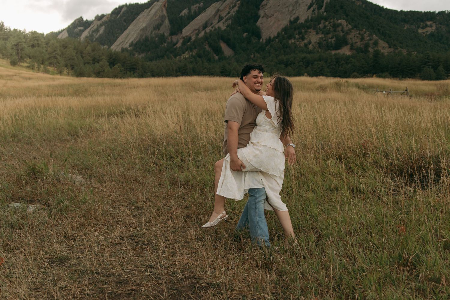 Couple embracing and spinning together in a golden grass field with mountain scenery behind them.