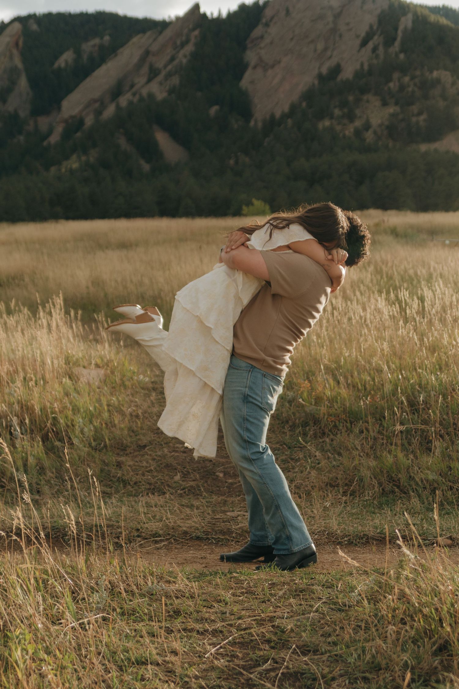Man lifts woman in white dress in a golden grass field with rocky mountain backdrop.