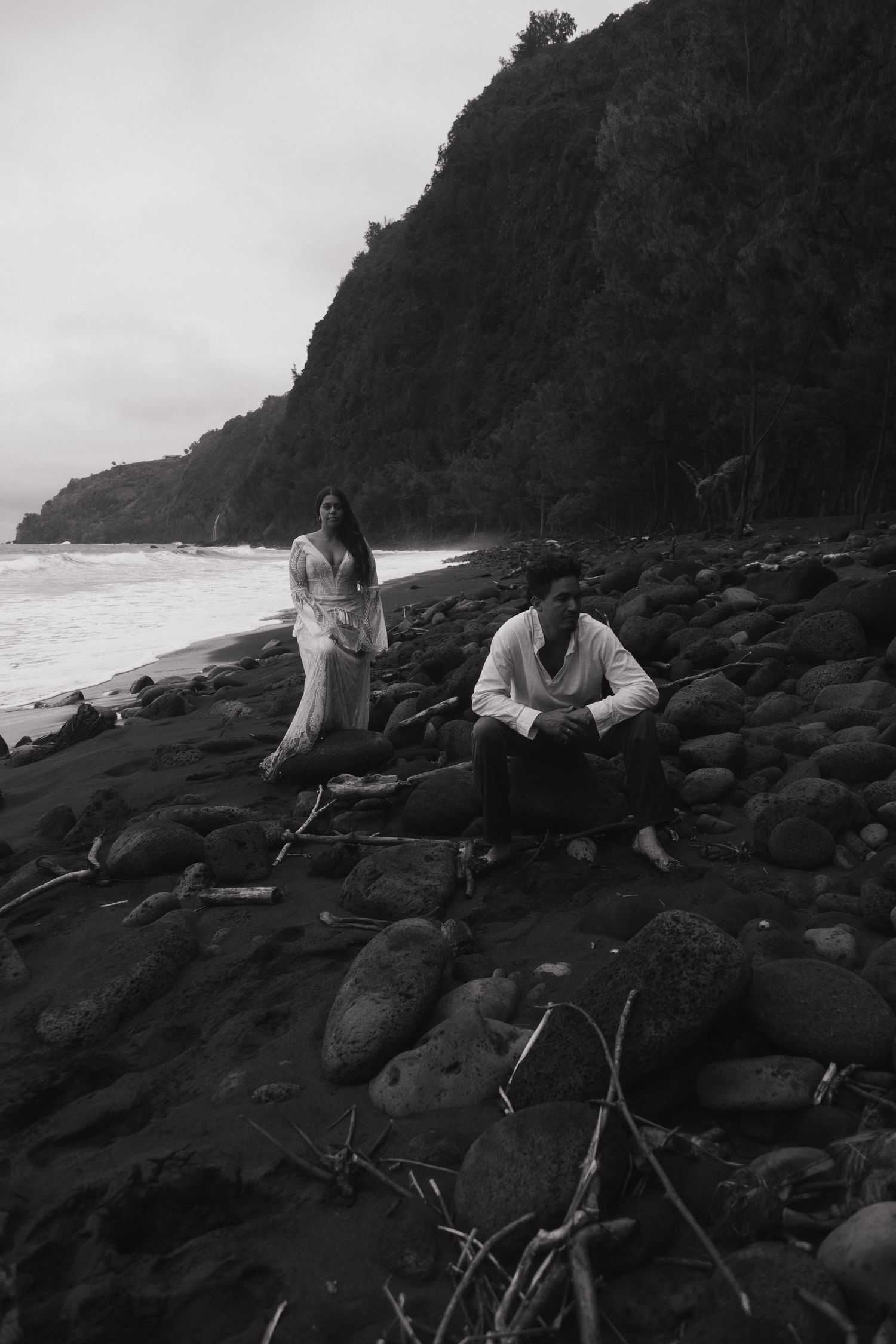 Black and white photo of couple sitting on rocky black sand beach with dramatic coastal cliffs behind them.