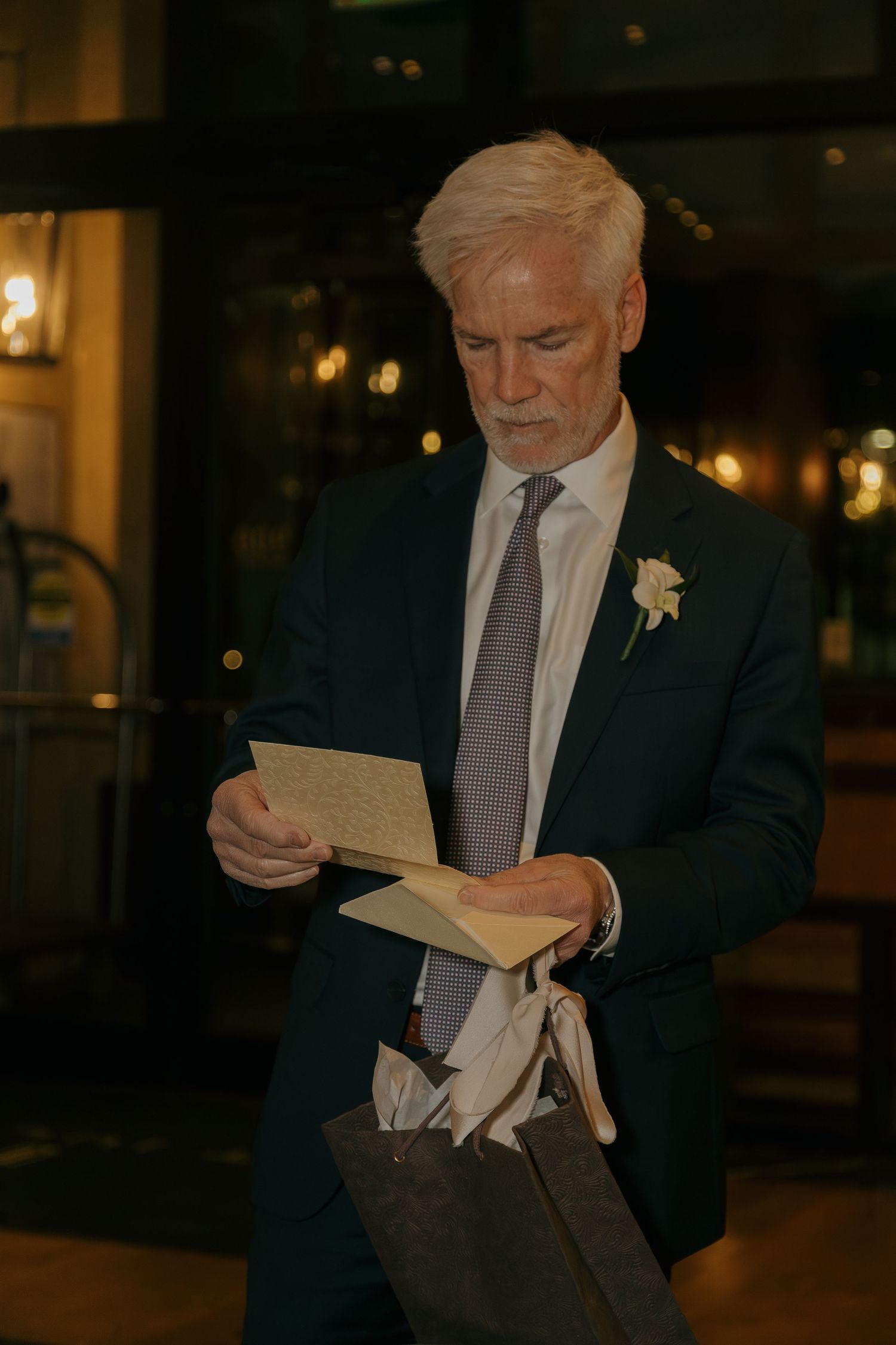 Elegant older man in dark suit reads a heartfelt card while holding a gift bag at a wedding venue.