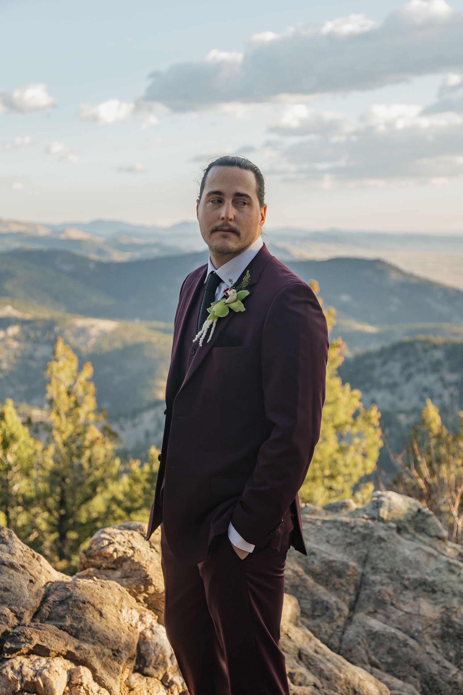 A groom in a burgundy suit with boutonniere posing on rocky mountain overlook.