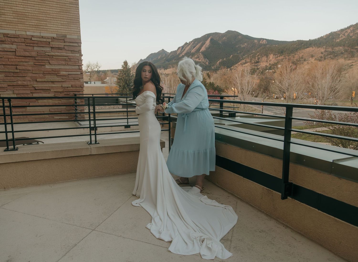 Bride in elegant white gown stands with elderly woman on balcony overlooking Colorado Flatirons.