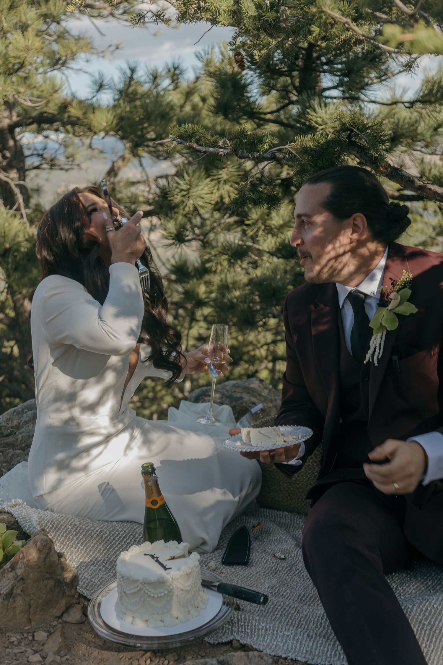 Bride and groom sharing wedding cake outdoors under pine trees with champagne during intimate elopement picnic.