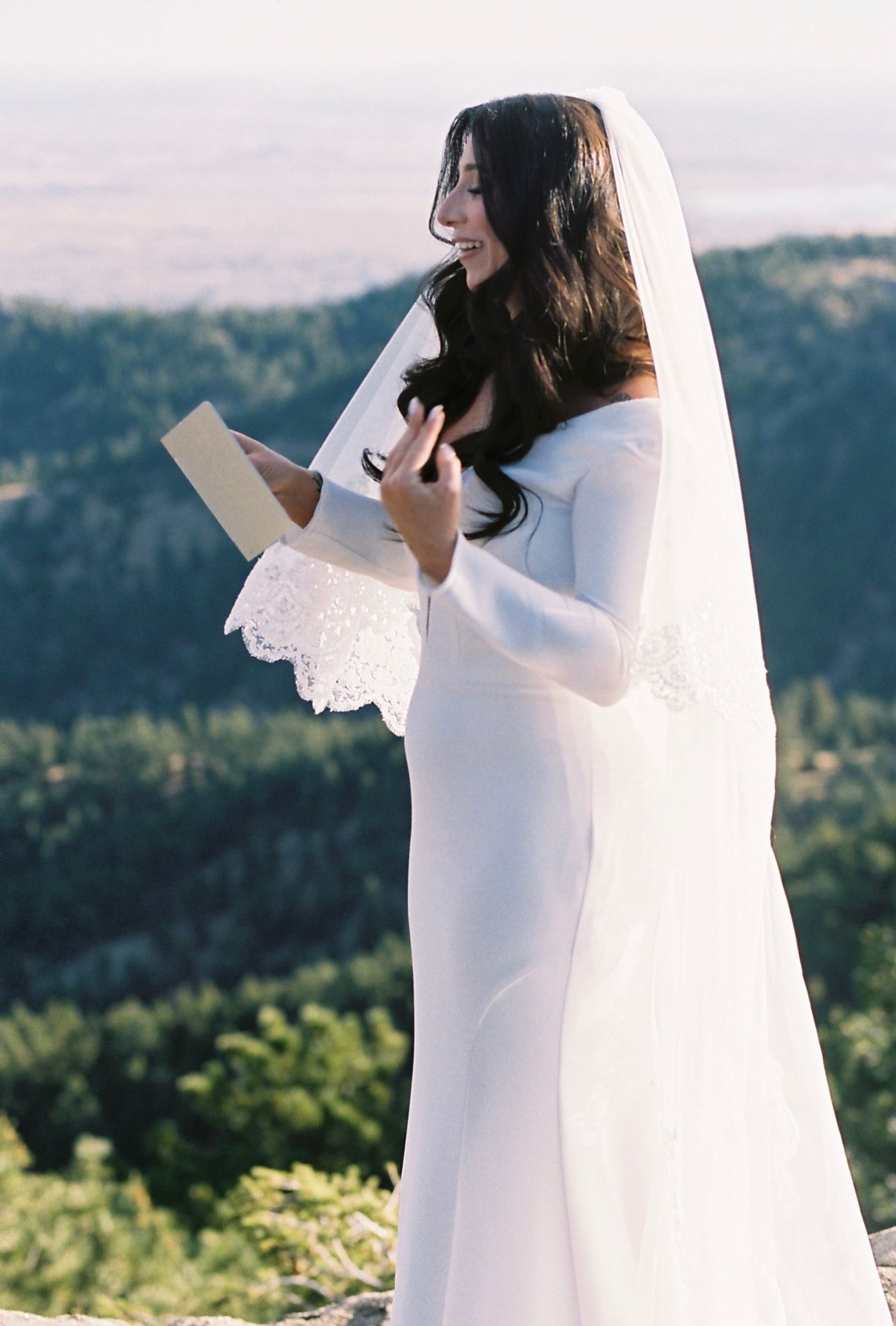 Bride in white gown and lace veil reads wedding vows with scenic mountain landscape behind her.