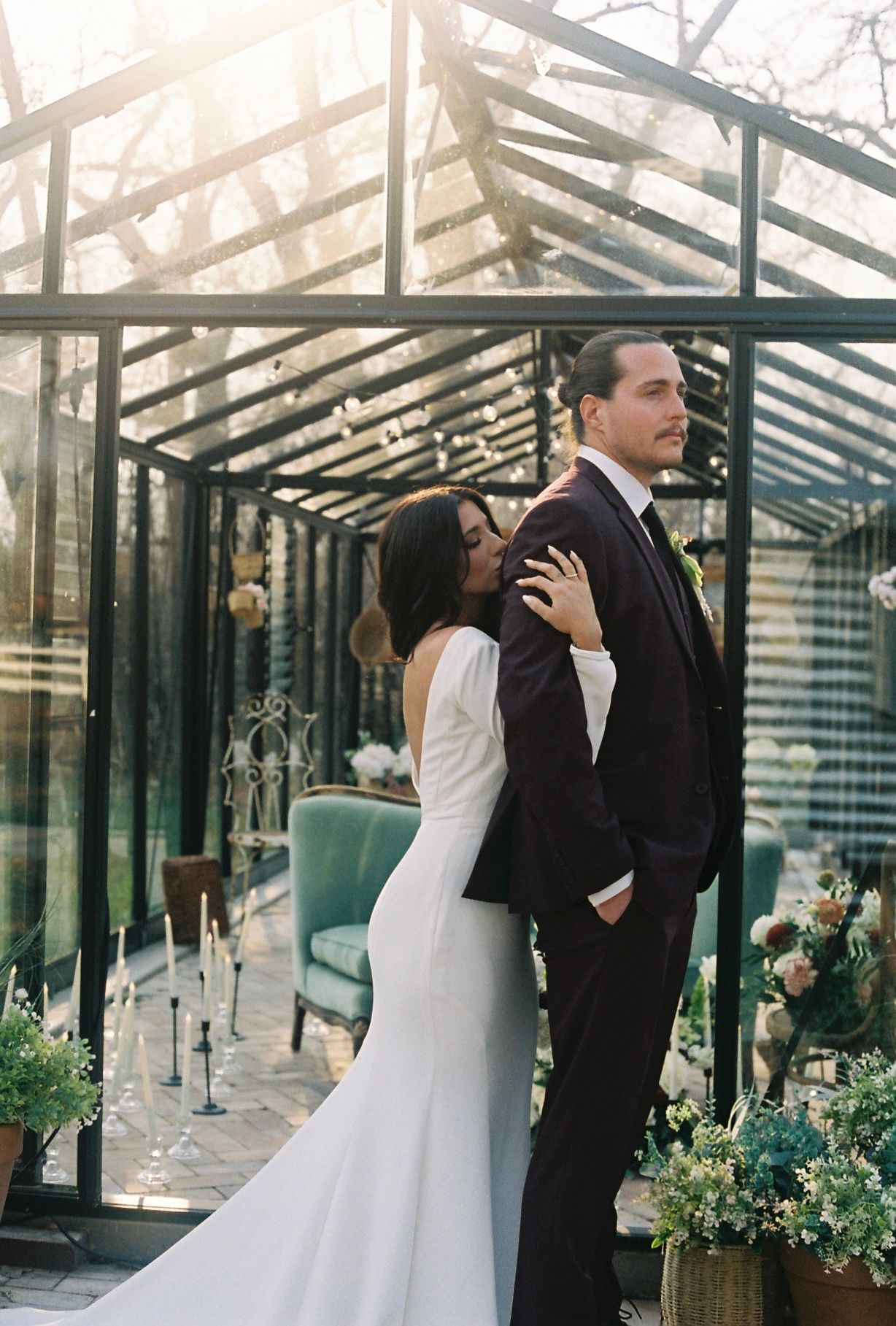 Bride embracing groom from behind near stunning glass greenhouse wedding venue in warm golden light.