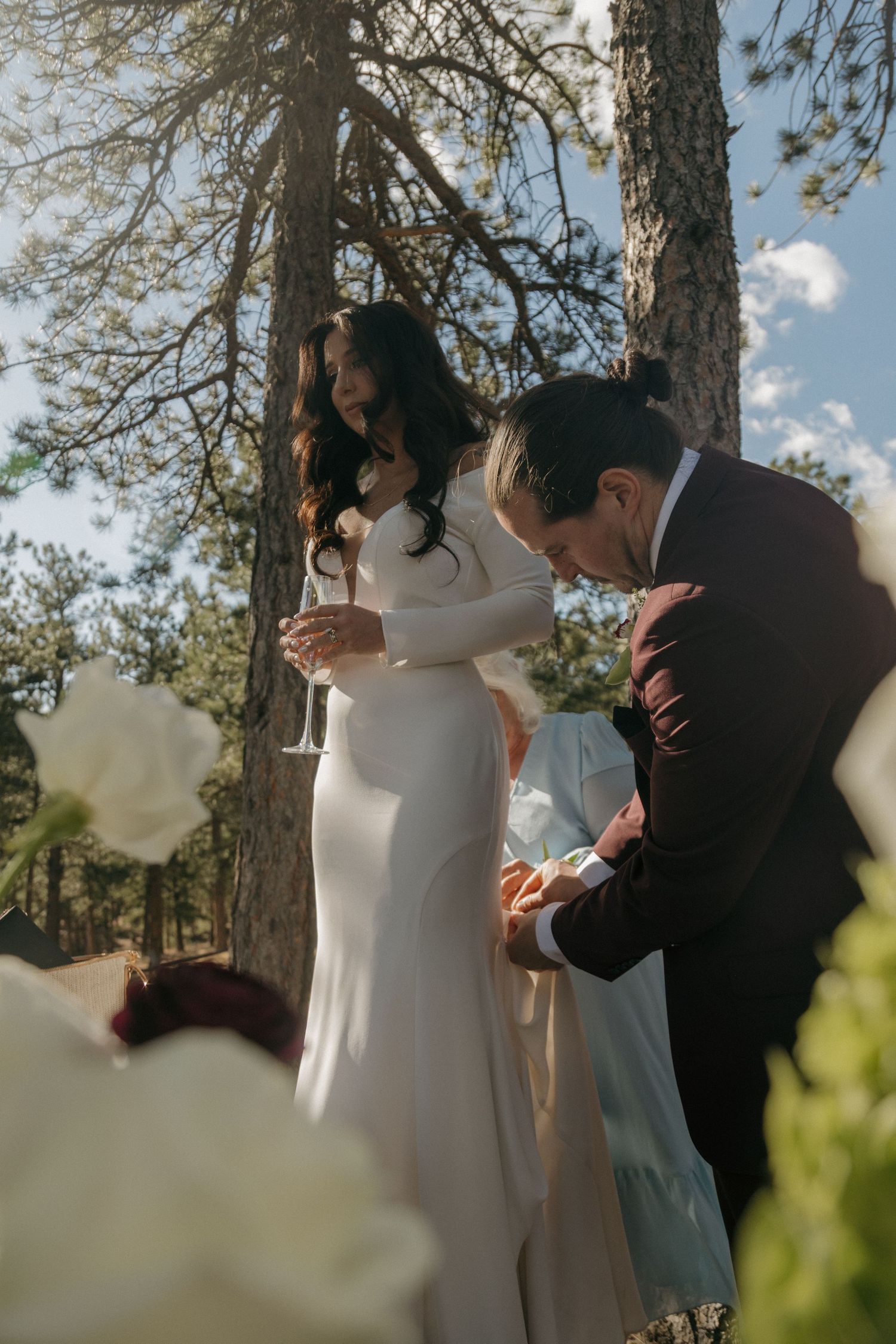 Groom in burgundy suit signs marriage license while bride holds champagne glass under pine trees outdoors.