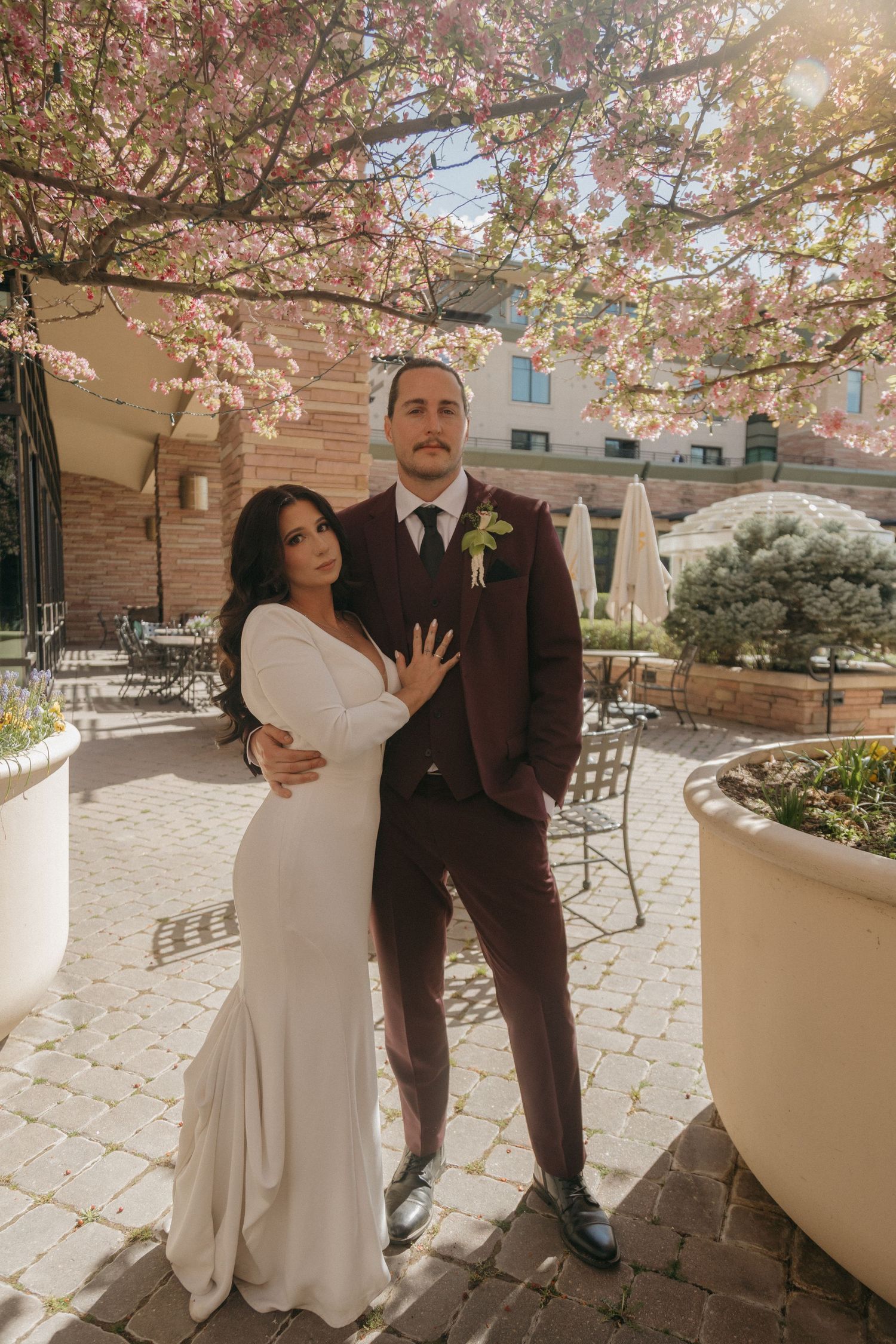 Elegant bride and groom posing together under blooming pink cherry blossom trees in a courtyard.