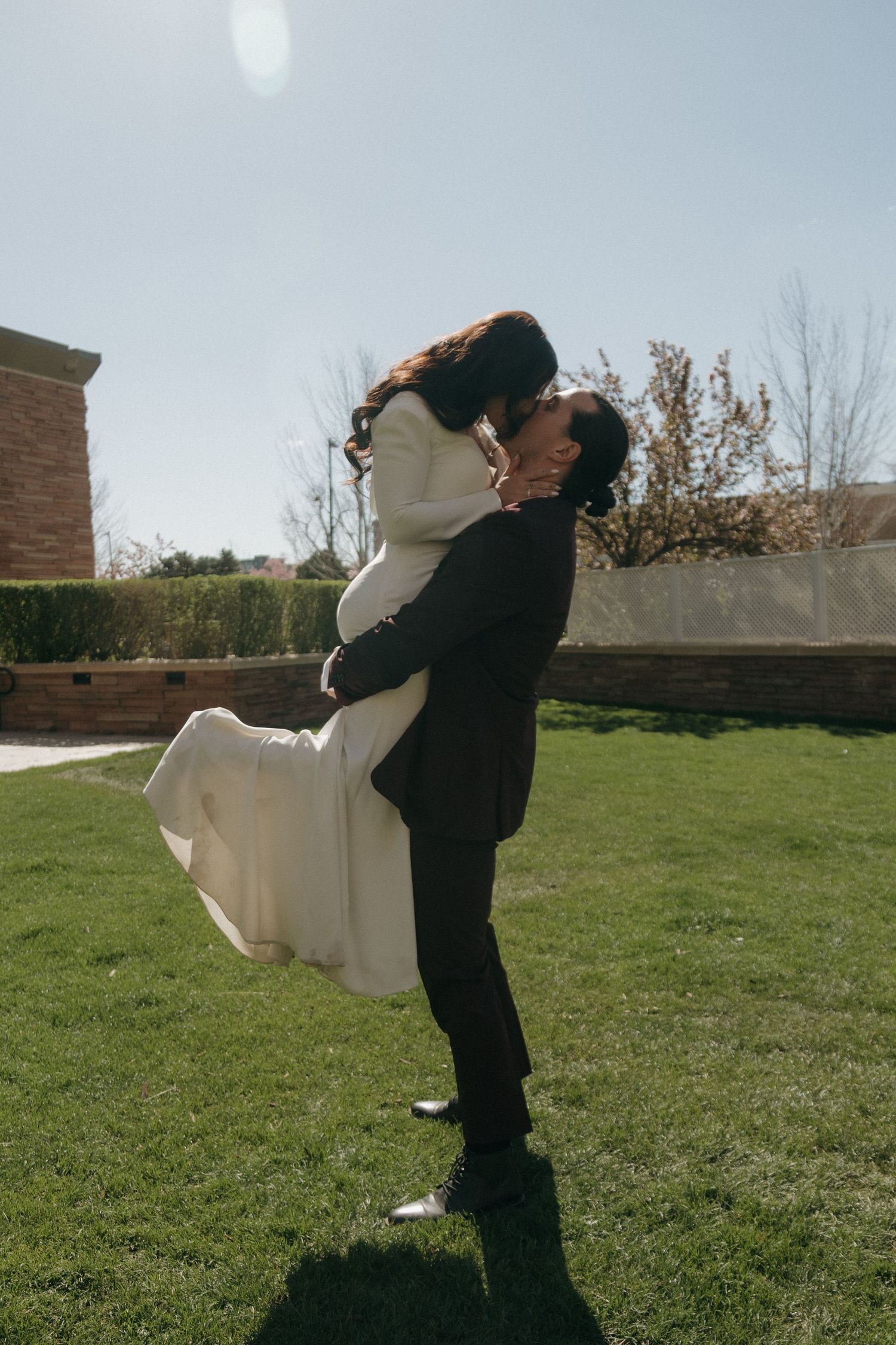Groom lifts bride in white wedding dress for a kiss on sunny green lawn during outdoor wedding celebration.
