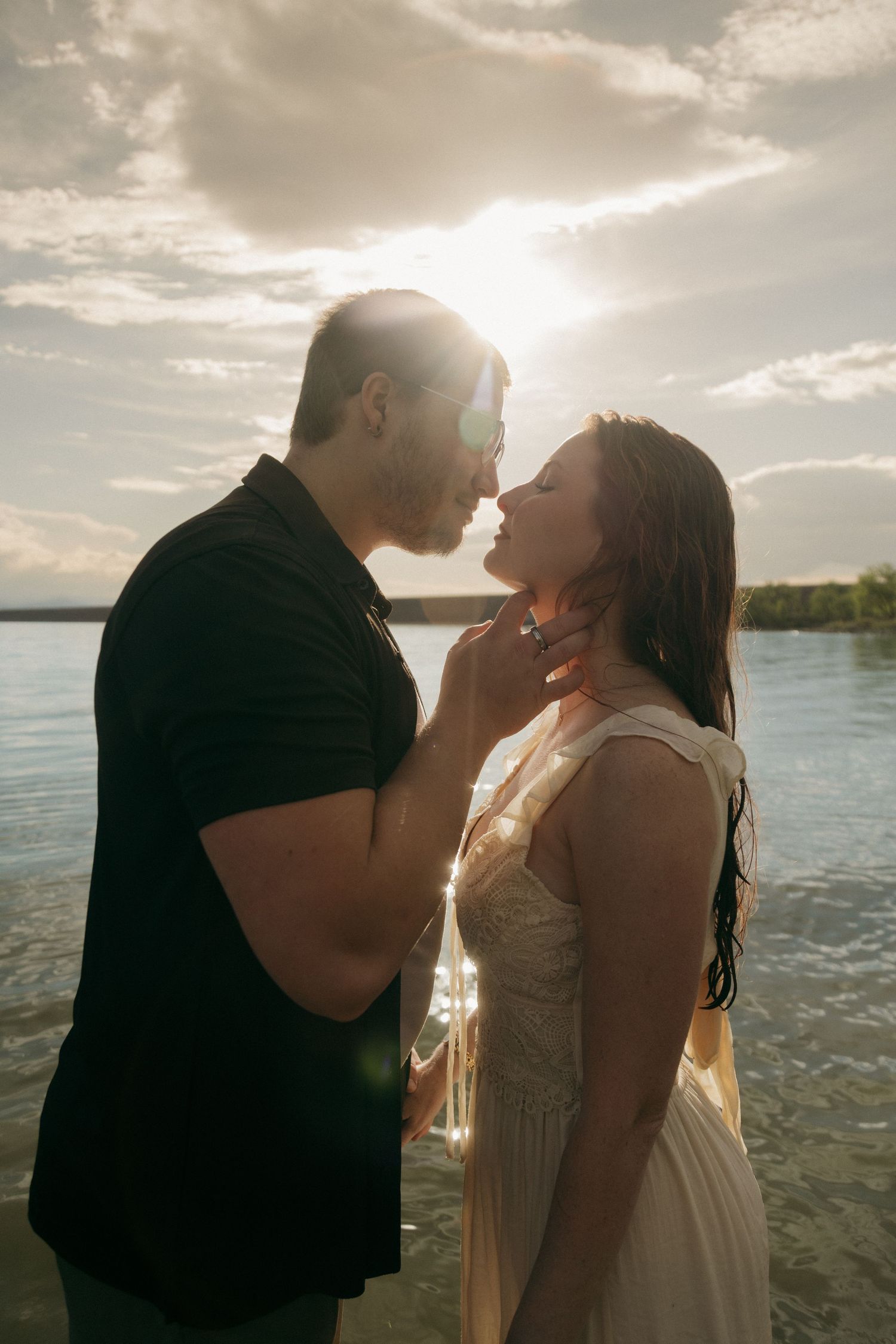 Romantic couple sharing a tender moment by a sunlit lake, man in black shirt gently touching woman's chin.