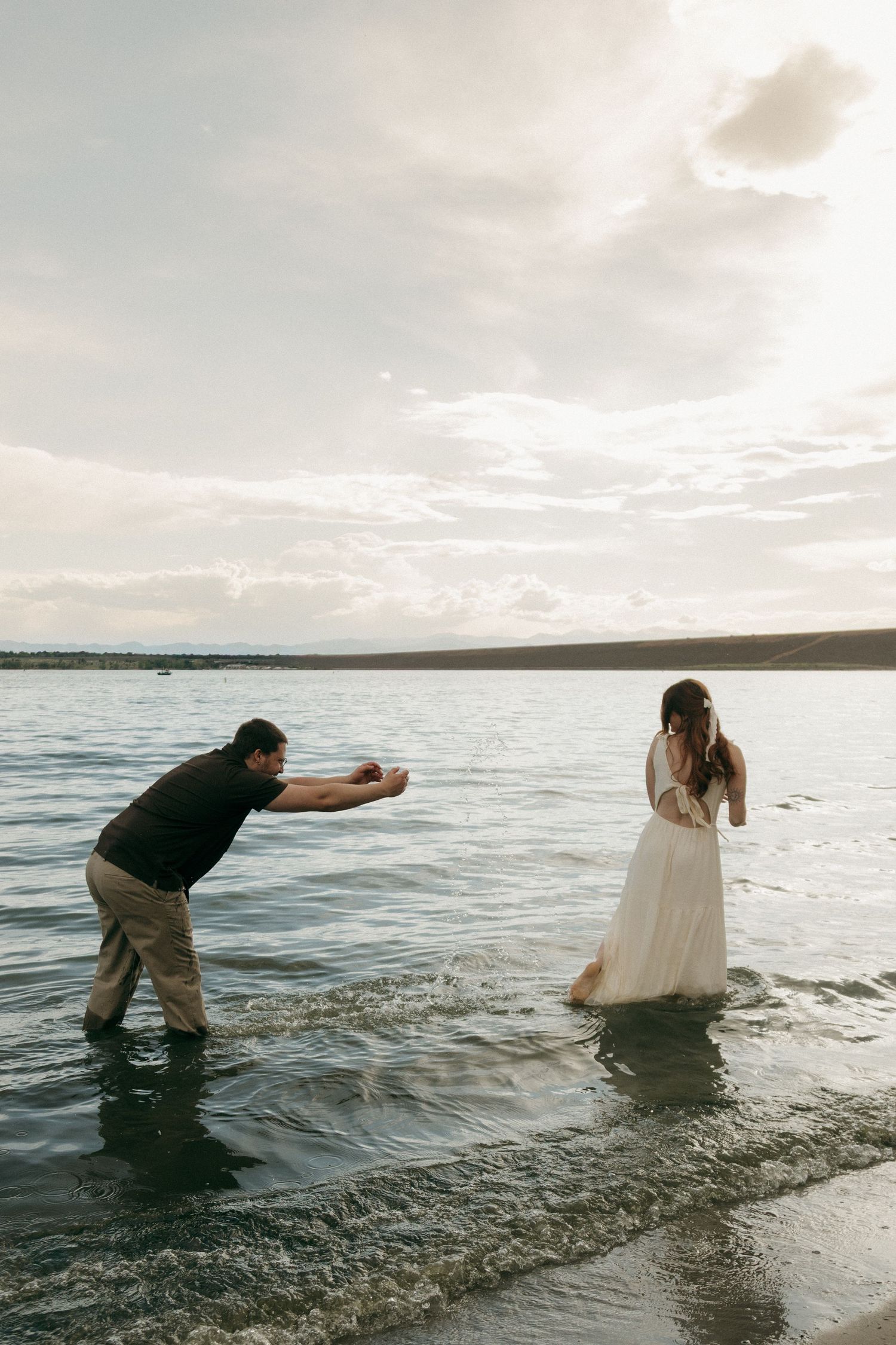 Man splashing water toward woman in white dress standing in a lake at sunset during engagement photo session.