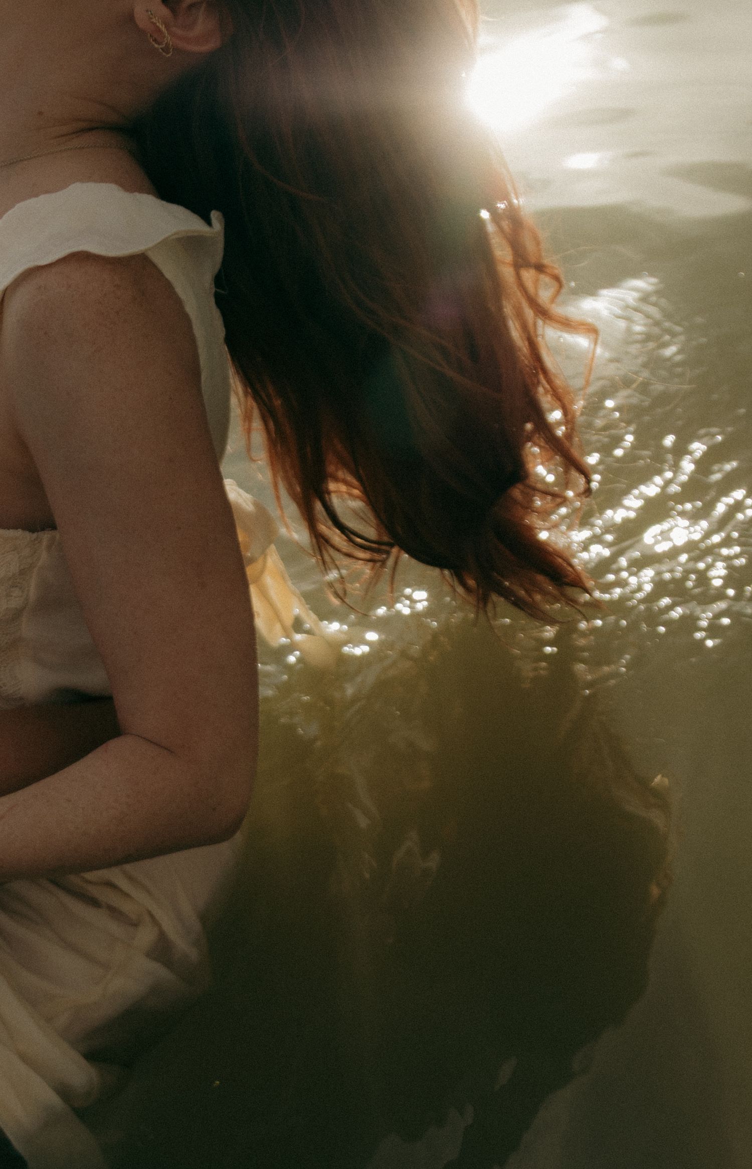 A woman in a white dress wades in sunlit water, her auburn hair catching the golden light.